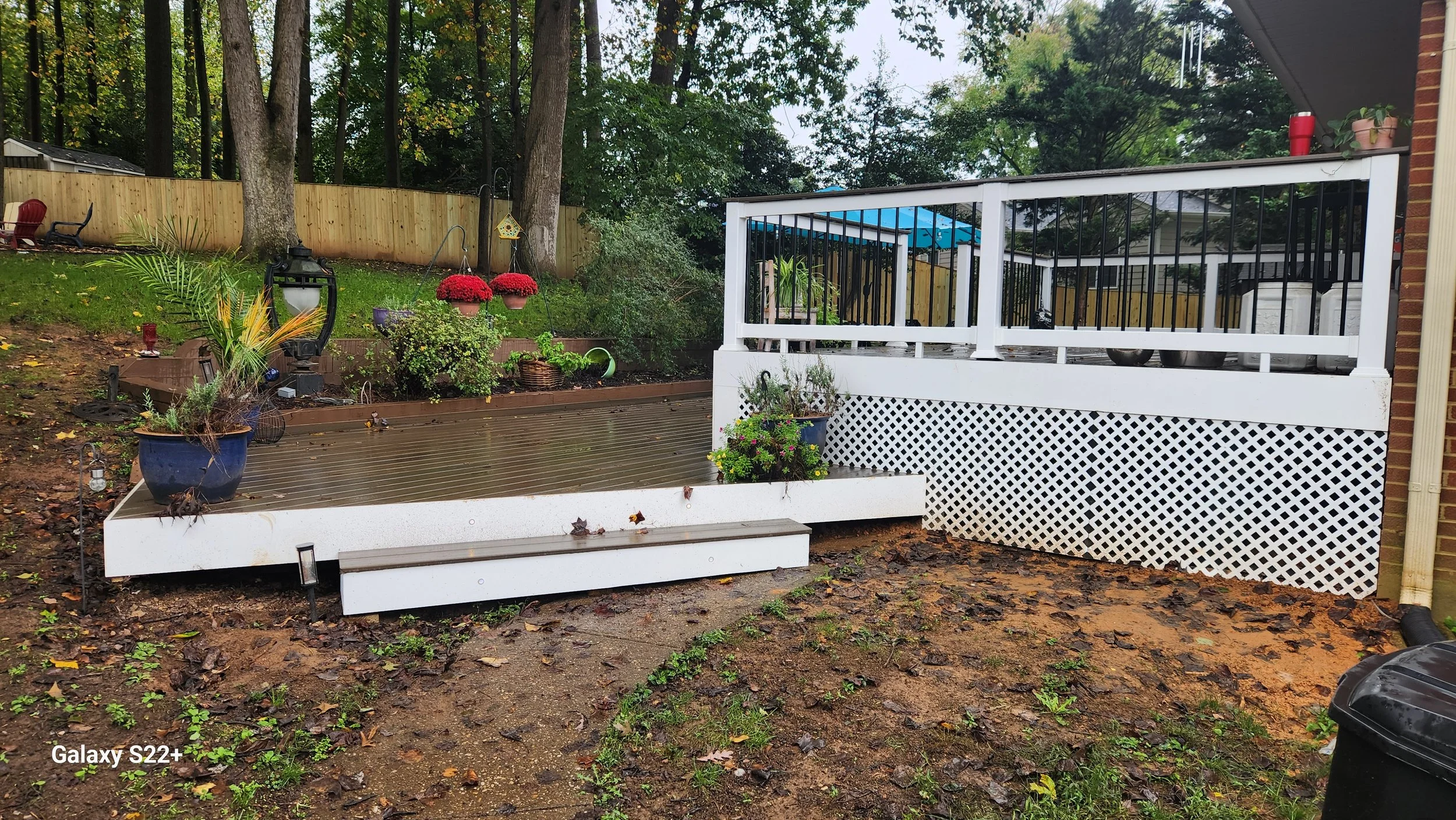 A backyard with a raised wooden deck with white framing and black railings, surrounded by trees, plants, and decorative flower pots, on a rainy day with wet ground.