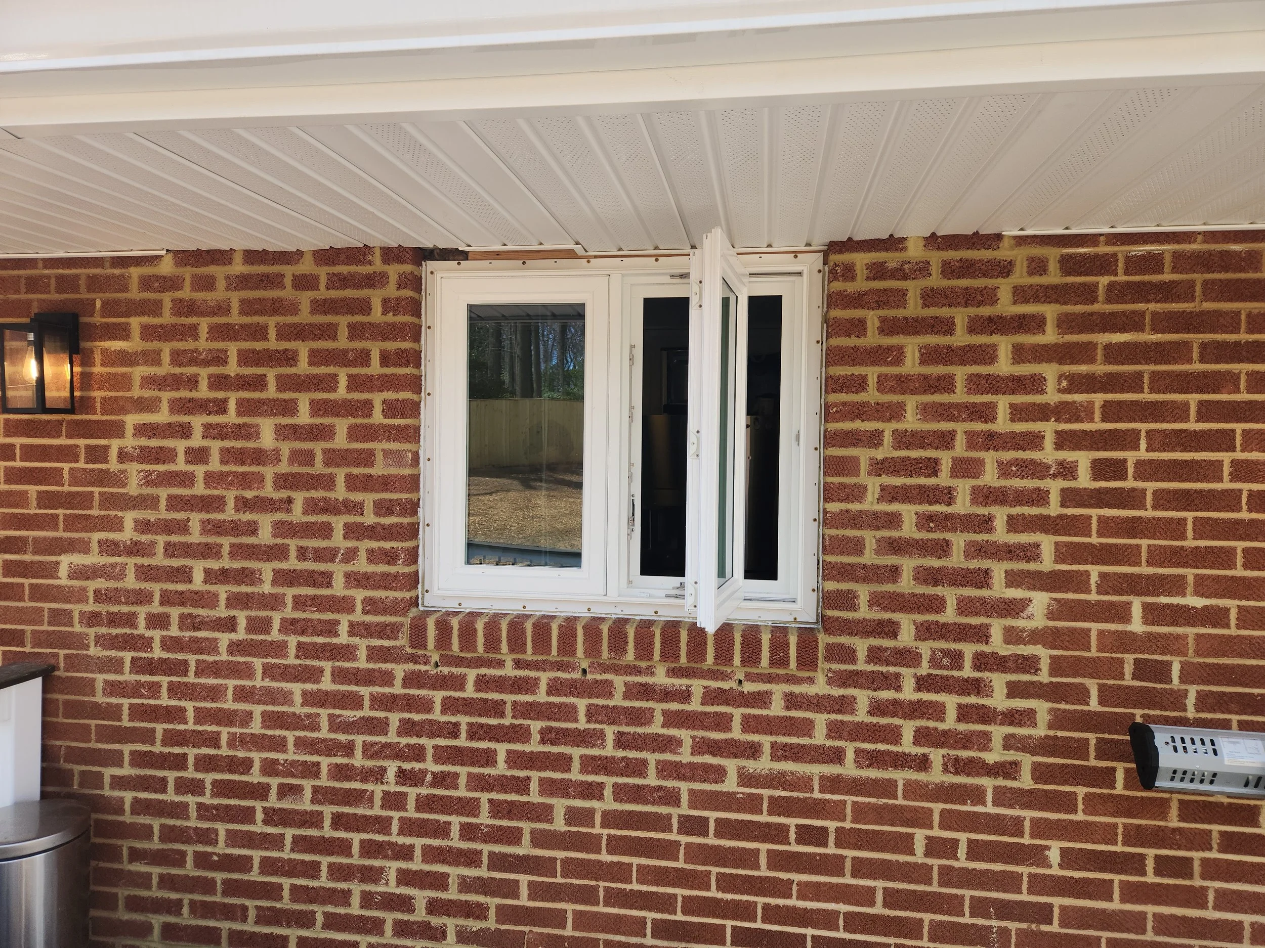 A white window on a red brick wall, with a half-open window panel. There is an outdoor wall light fixture on the left and a small white object on the right.