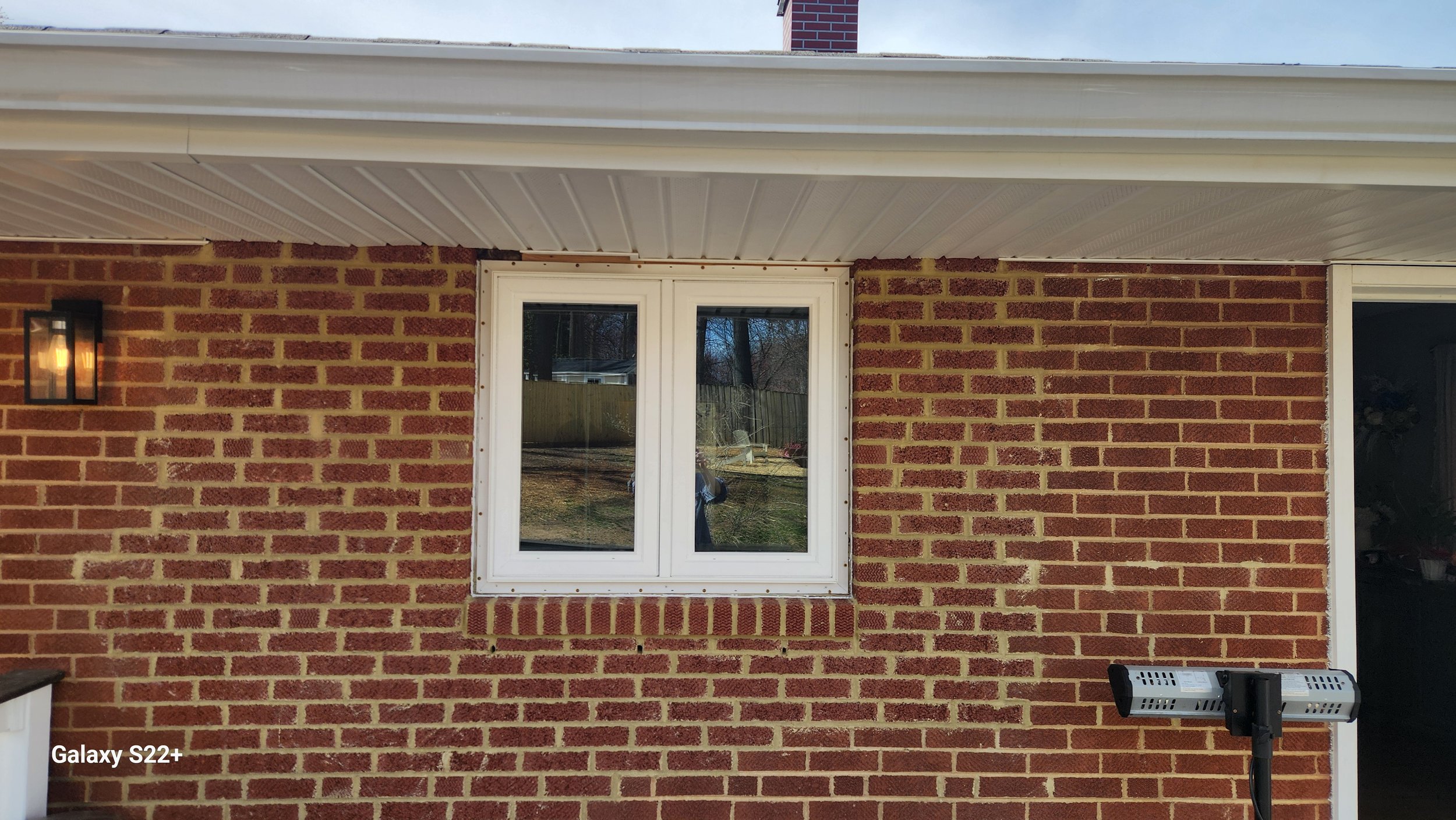 View of the exterior brick wall of a house with a white-framed window, outdoor wall sconce, and a mounted outdoor heater or light fixture, with a backyard visible through the window.