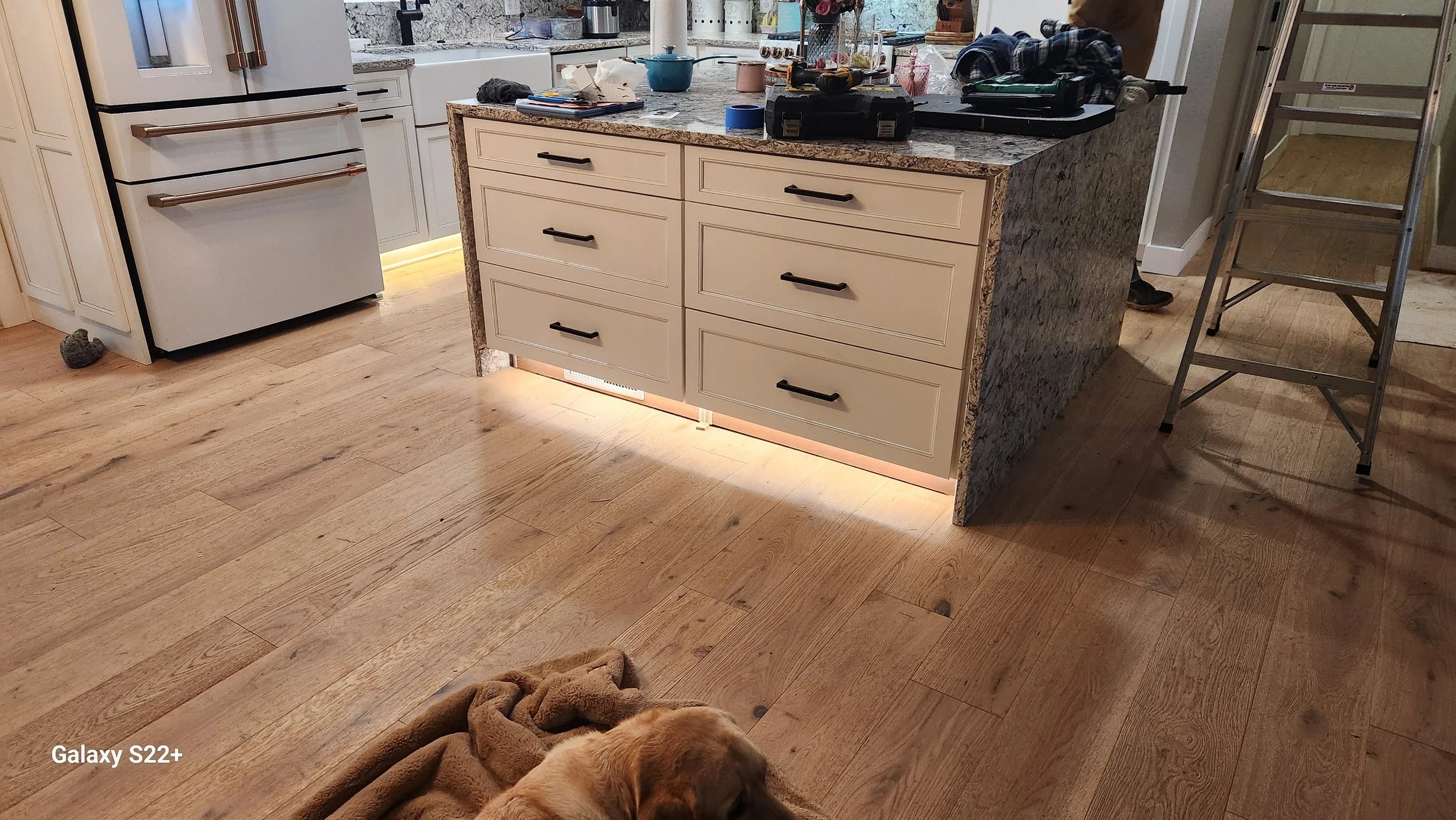 A modern kitchen with wooden flooring, white cabinets, and a granite kitchen island with drawers. There are various items on the island, including tools and household items. To the right, a ladder leans against the wall. A puppy rests on a blanket ne