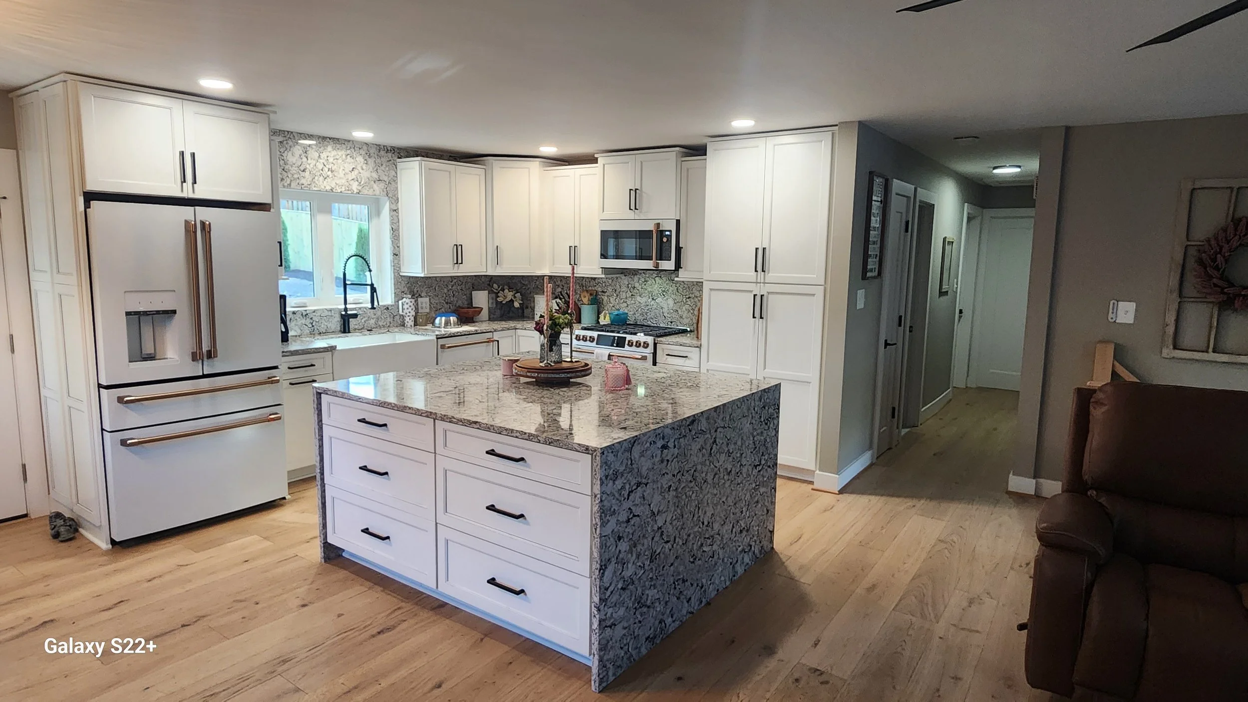 Modern kitchen with white cabinets, granite countertop, and a large island in the center. Includes a white refrigerator, microwave, and stove. There's a window above the sink, and the floor is light wood.