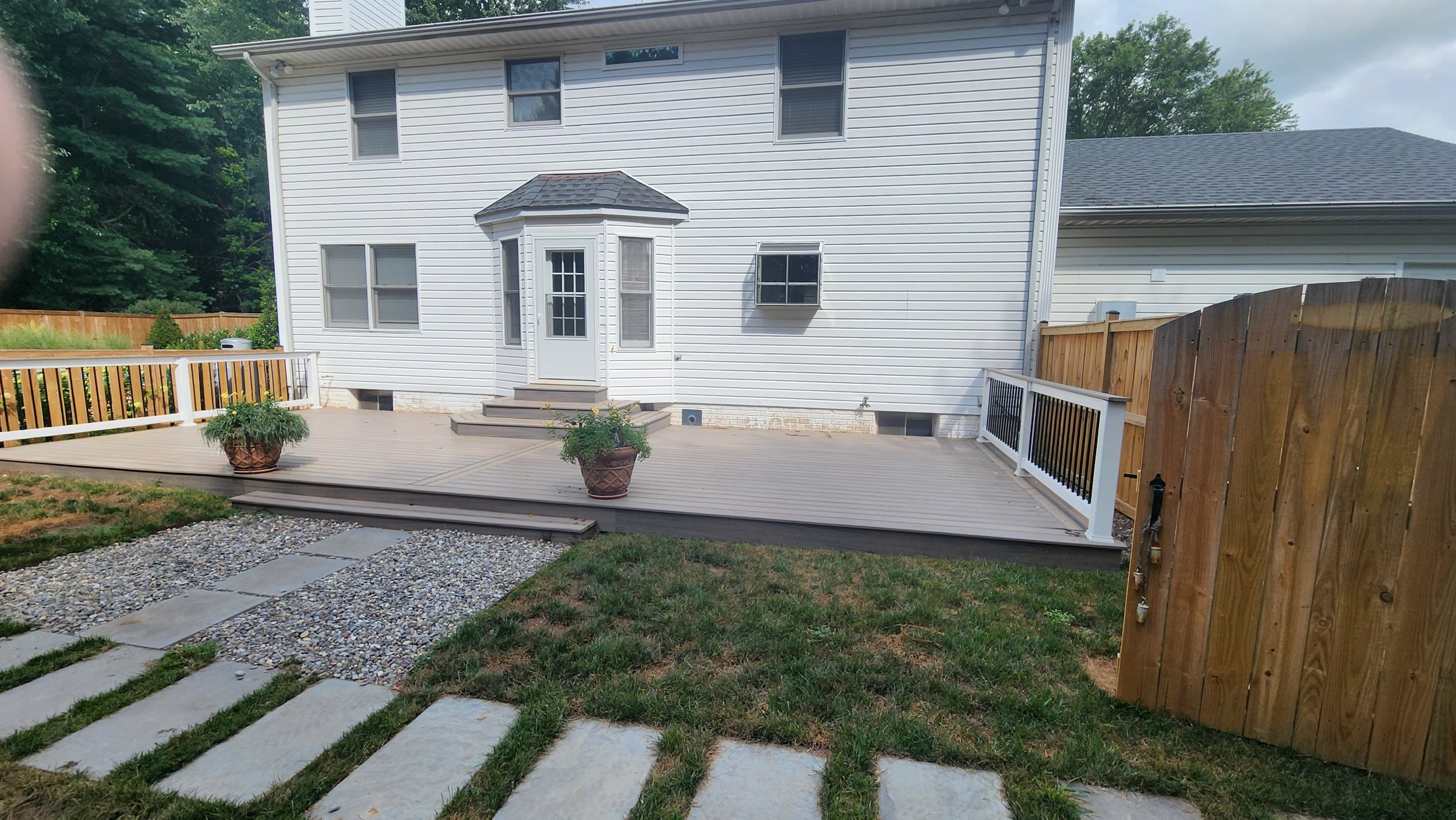 Backyard with a raised wooden deck, two large potted plants, a gravel pathway, a grassy lawn, and a white house with a small bay window and stairs leading to a door.