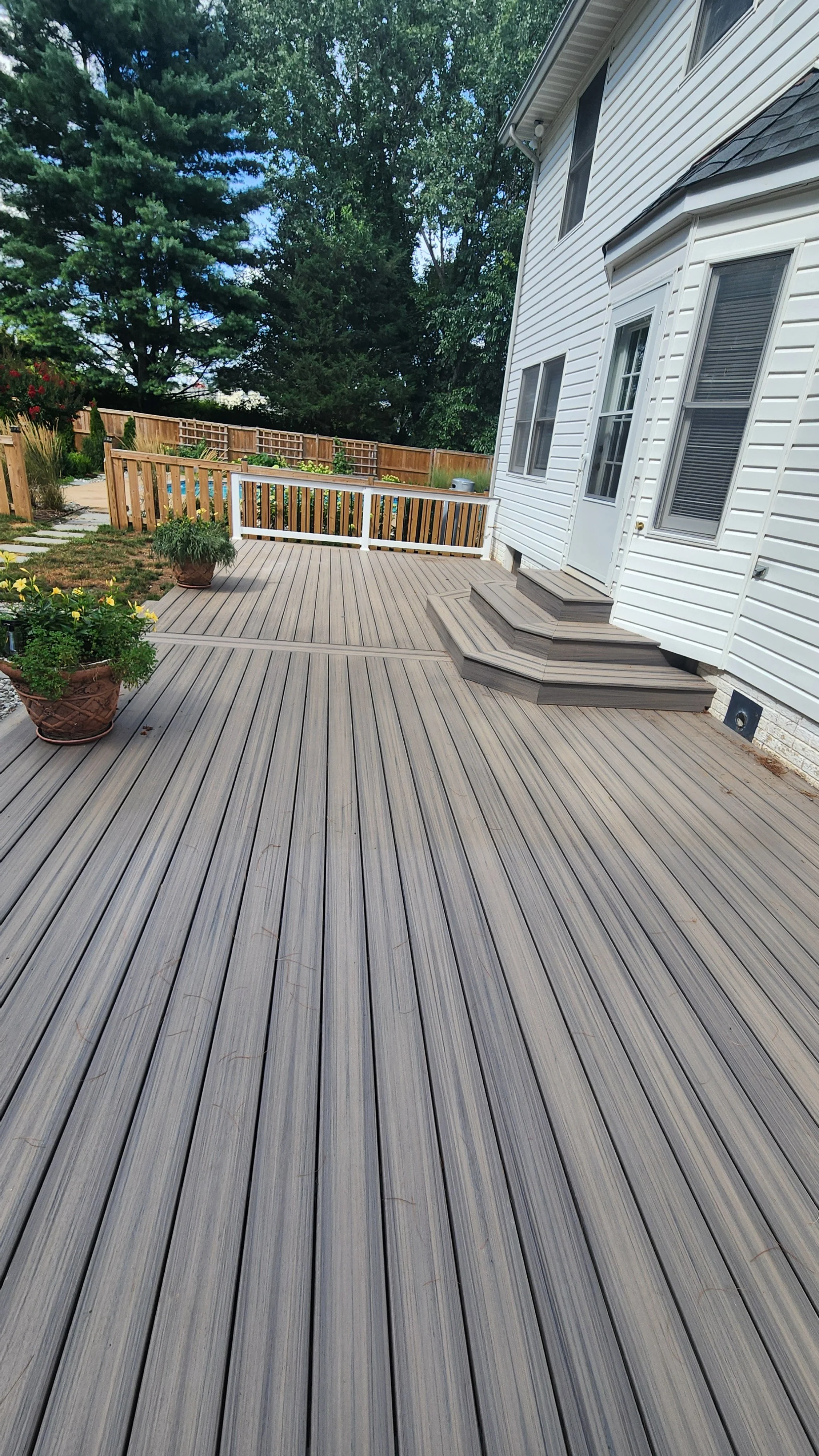 A spacious backyard deck made of gray composite wood planks attached to a white house with a white door and multiple windows, surrounded by a new wooden fence, some potted plants, and lush green trees in the background.