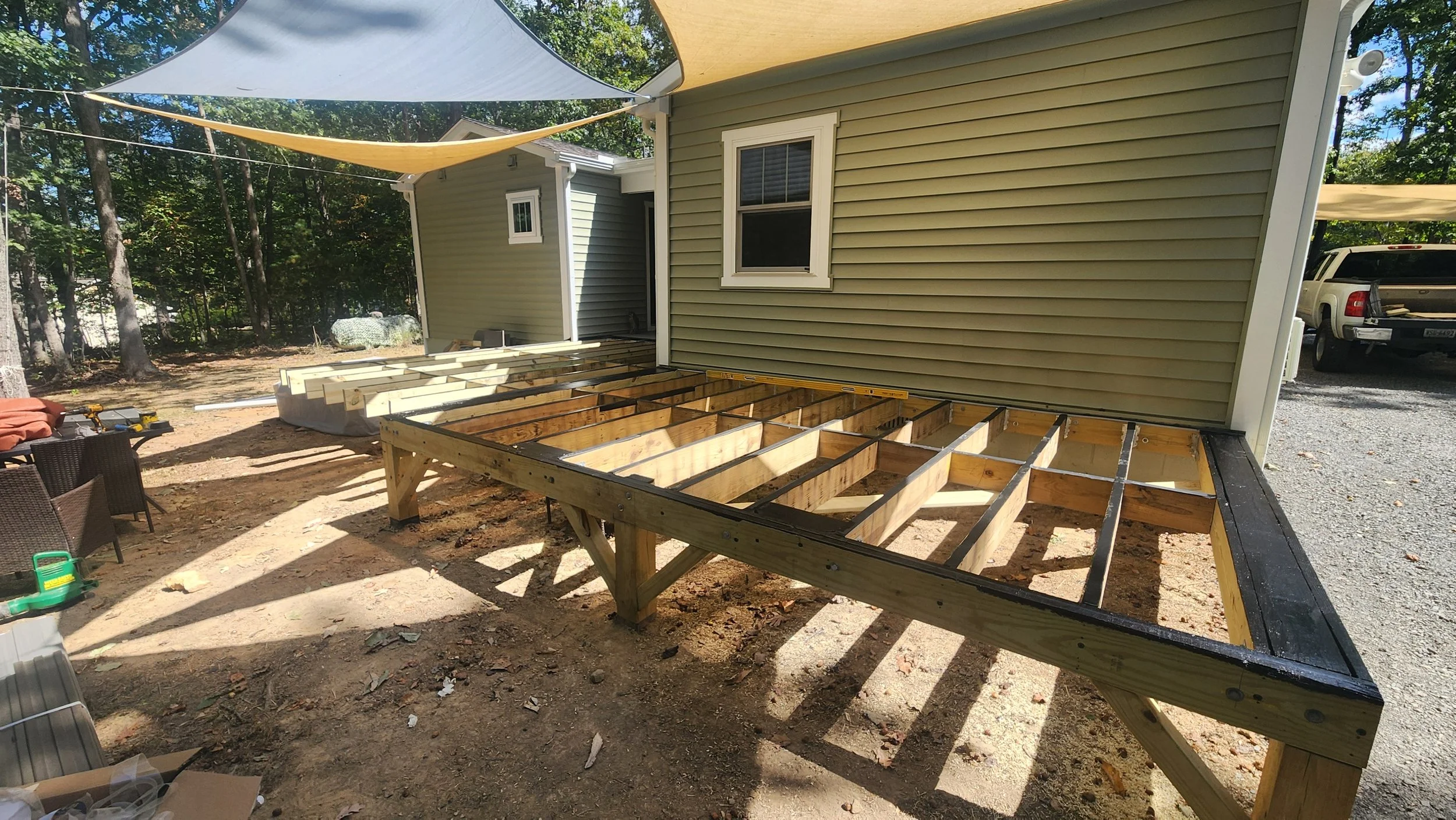 Wooden deck frame under construction outside a house, with shade sails overhead and parked vehicles in the background.