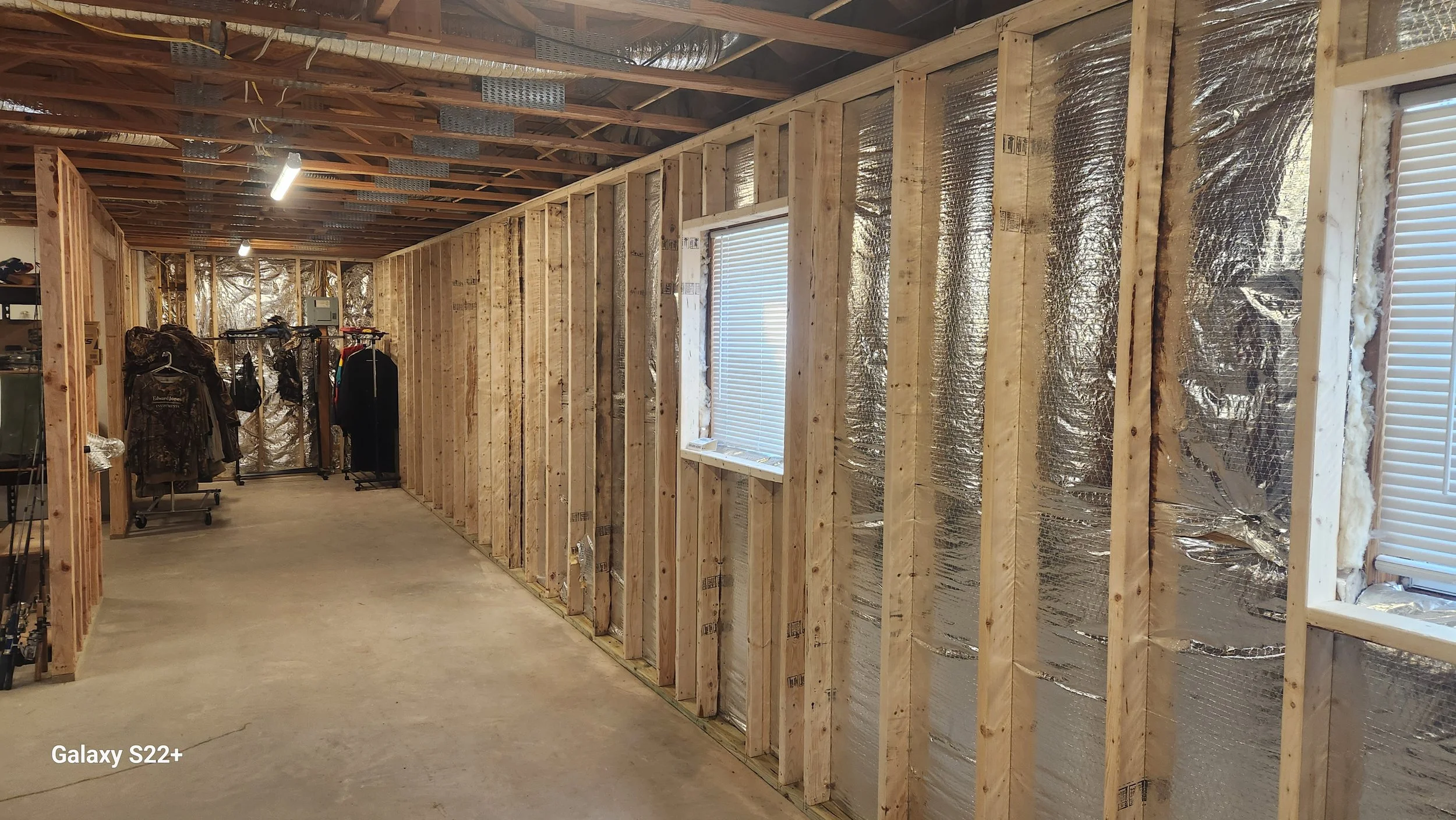 Unfinished interior of a house under construction with exposed wooden framing, insulation, and small clothing racks with jackets in the background.