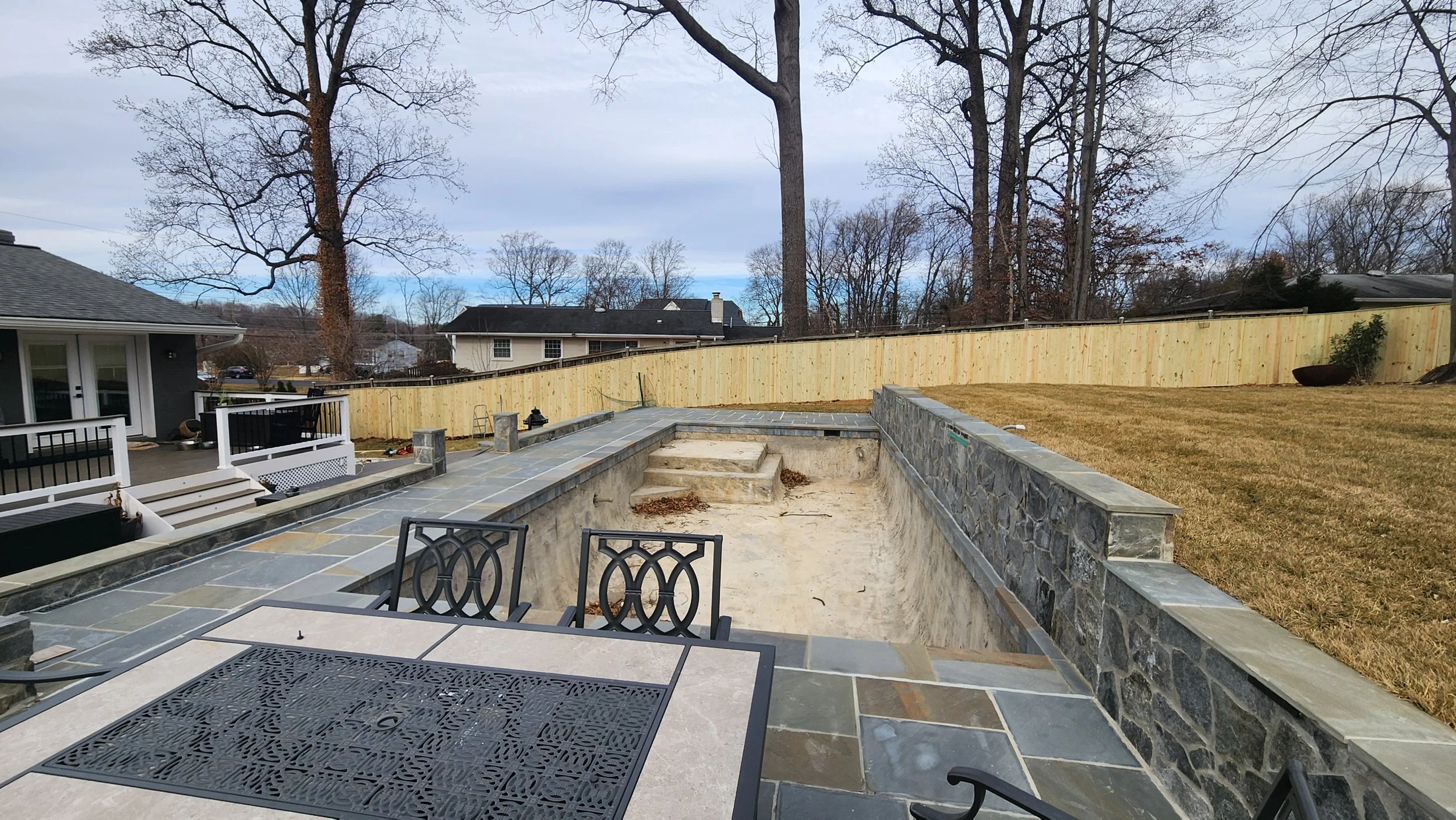 Backyard with an in-ground swimming pool under construction, surrounded by stone patio and a wooden privacy fence, with patio furniture, a tree on the left, and neighboring houses in the background.