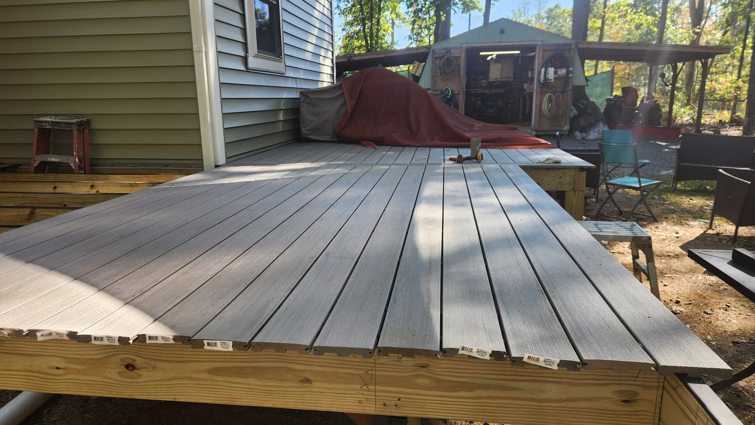 A partially completed wooden deck with light gray composite planks, a power drill on the deck, and a covered object on the side. In the background, there is a shed with tools and garden equipment, and outdoor furniture including chairs and a small ta