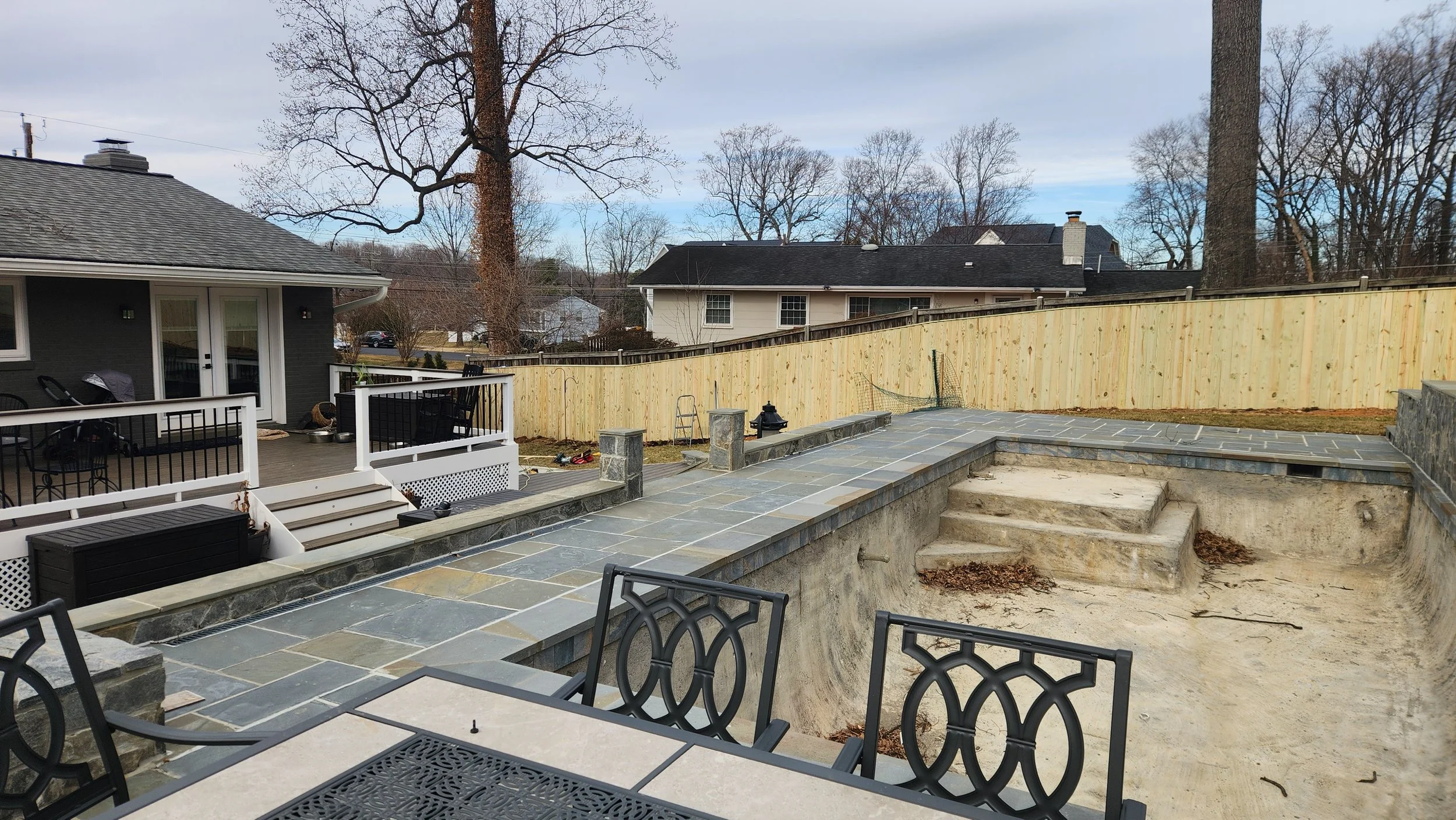 Backyard under renovation with a partially built stone patio, steps, and empty swimming pool area, surrounded by a wooden fence and neighboring houses.