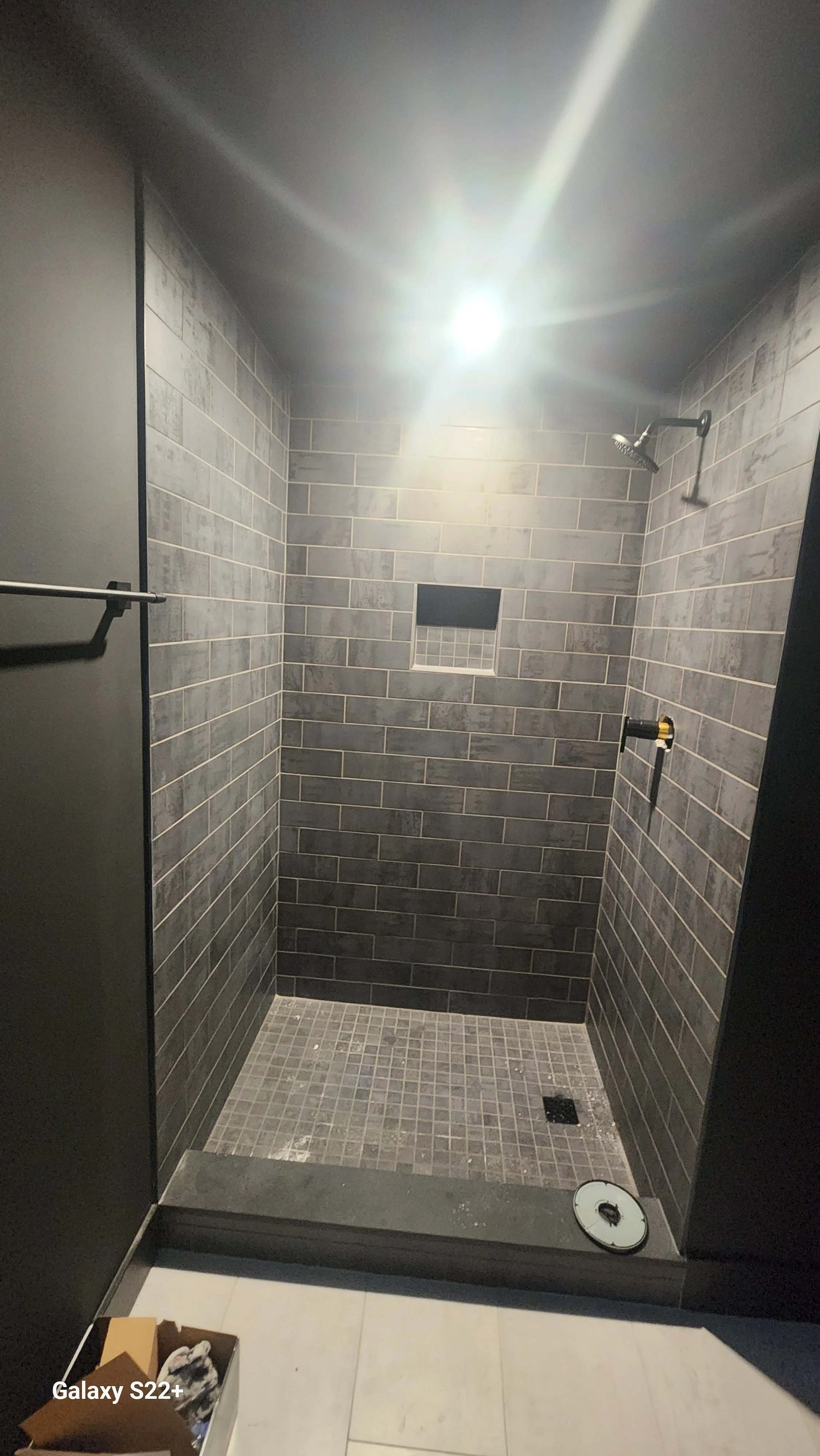 A shower area with dark grey subway tile walls, a niche, a showerhead, and a floor drain, with construction items in the foreground.