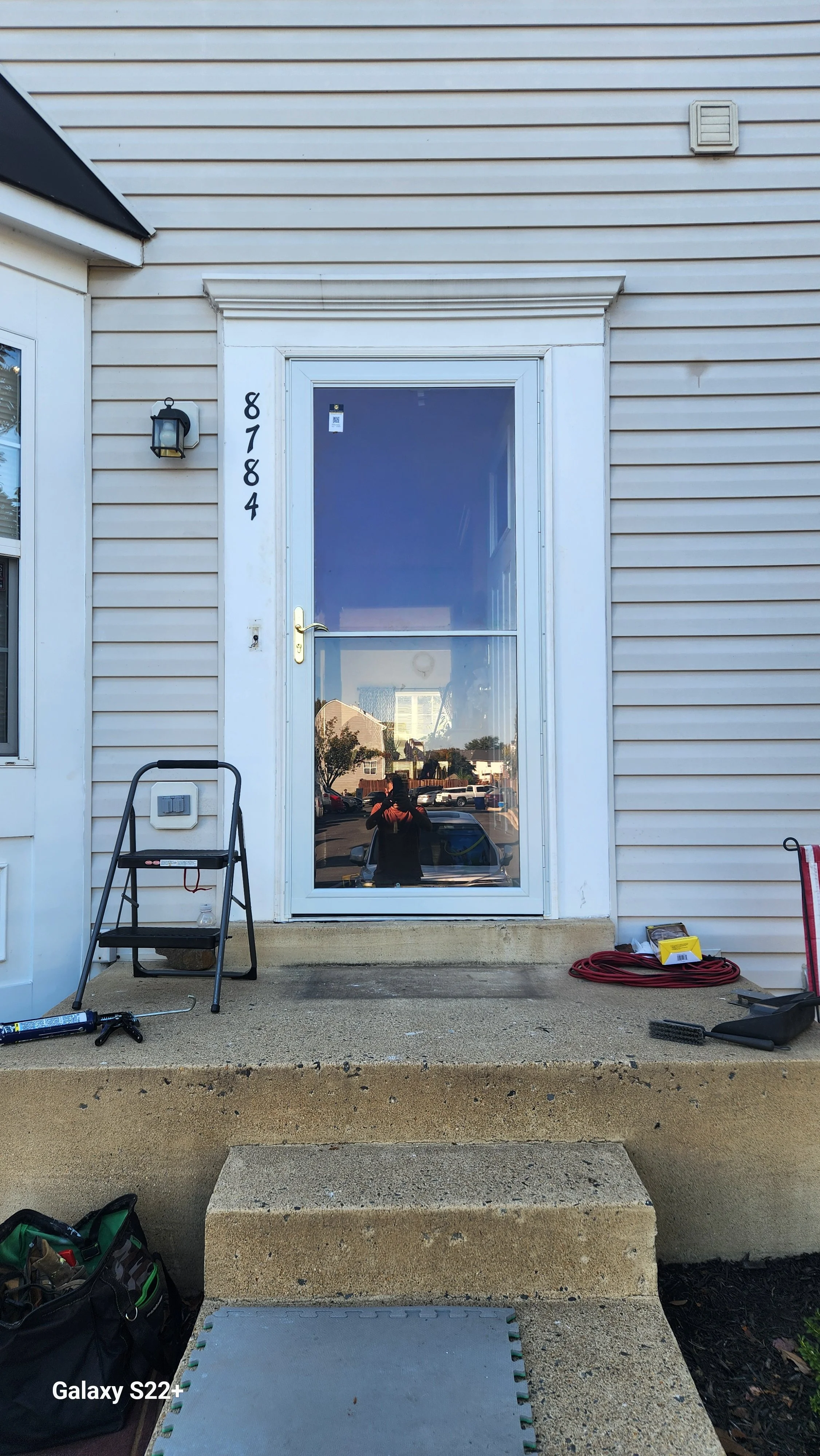Front door of a house with the house number 8784 on the wall. The door has a large glass pane that reflects the surrounding houses and cars. There is a black outdoor light fixture to the left of the door. The steps and porch have various tools and eq