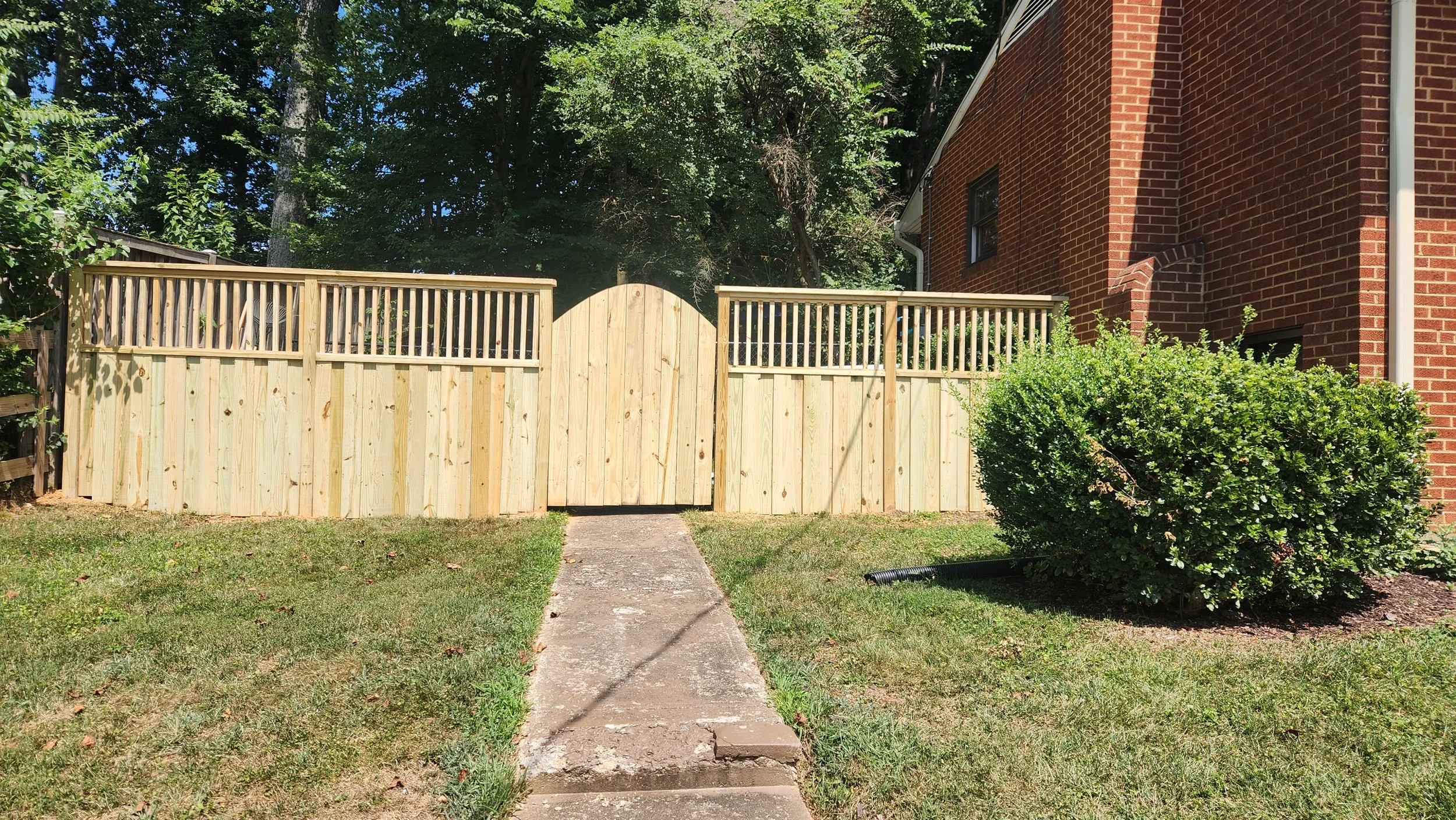 Front yard with a new wooden fence, a concrete pathway, green grass, a bush, and a red brick house.
