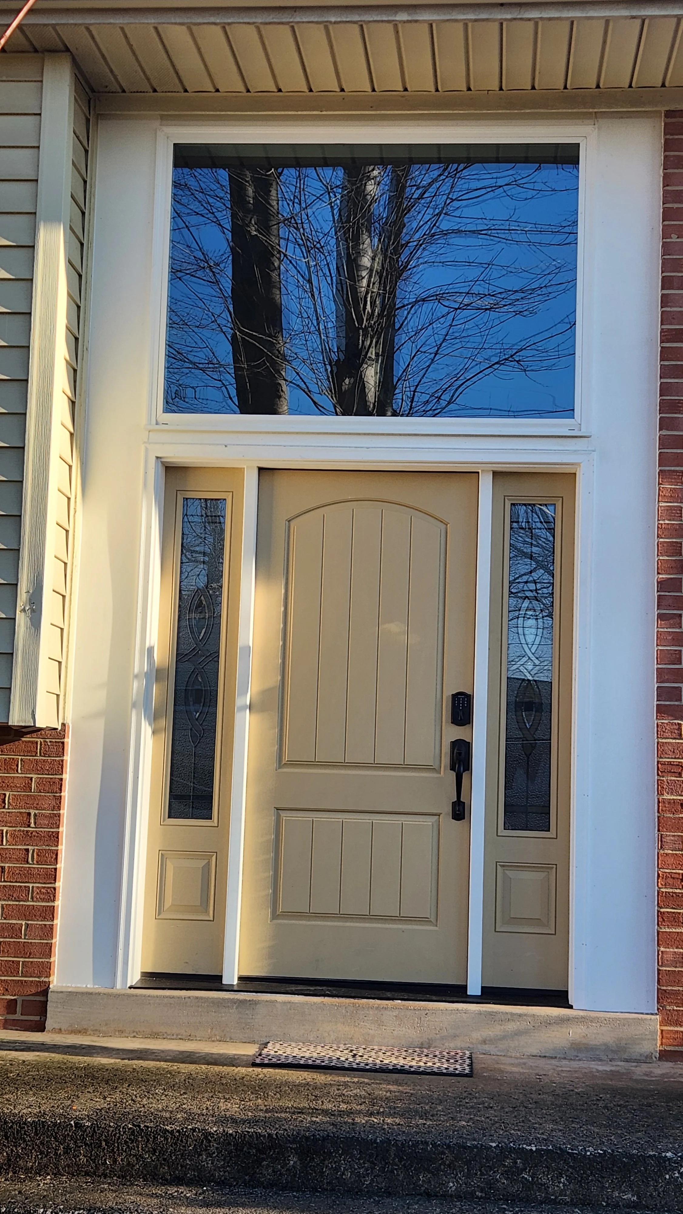 Front door of a house with a large window above, beige door with vertical design, and sidelights with decorative glass, set against brick and siding exterior with a concrete porch and mat.