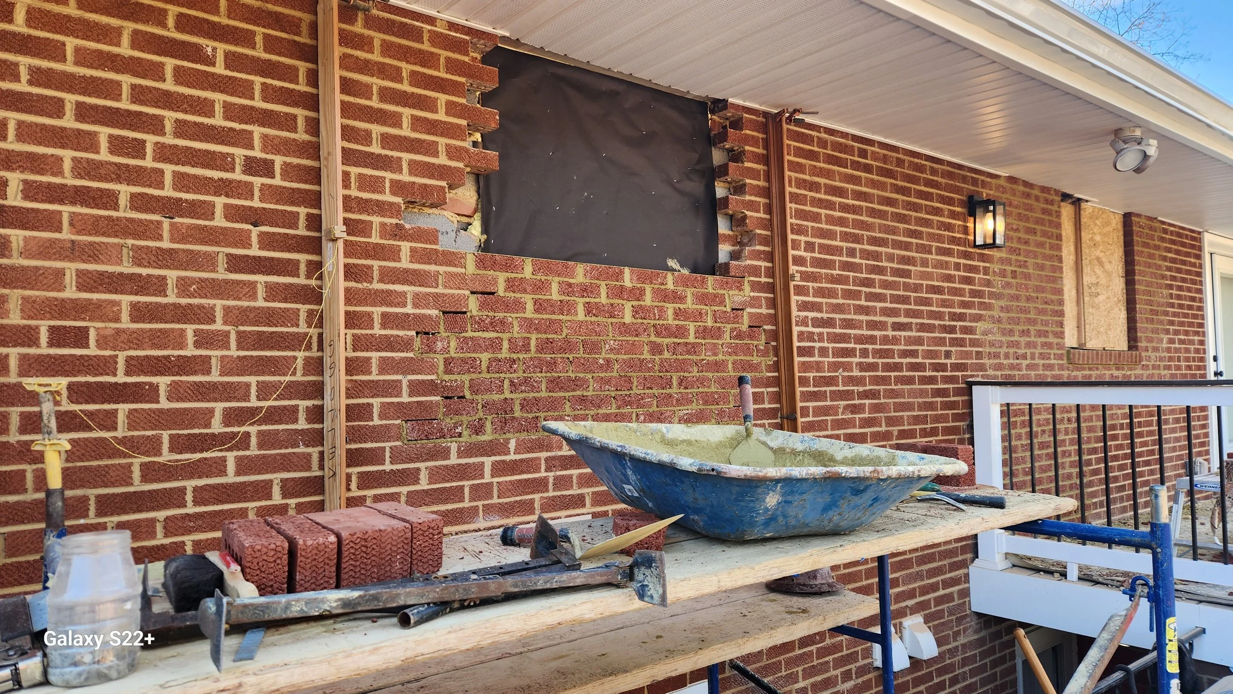 A brick house porch under renovation showing an unfinished wall, construction tools, bricks, a cement tub, and a worker's scaffolding.