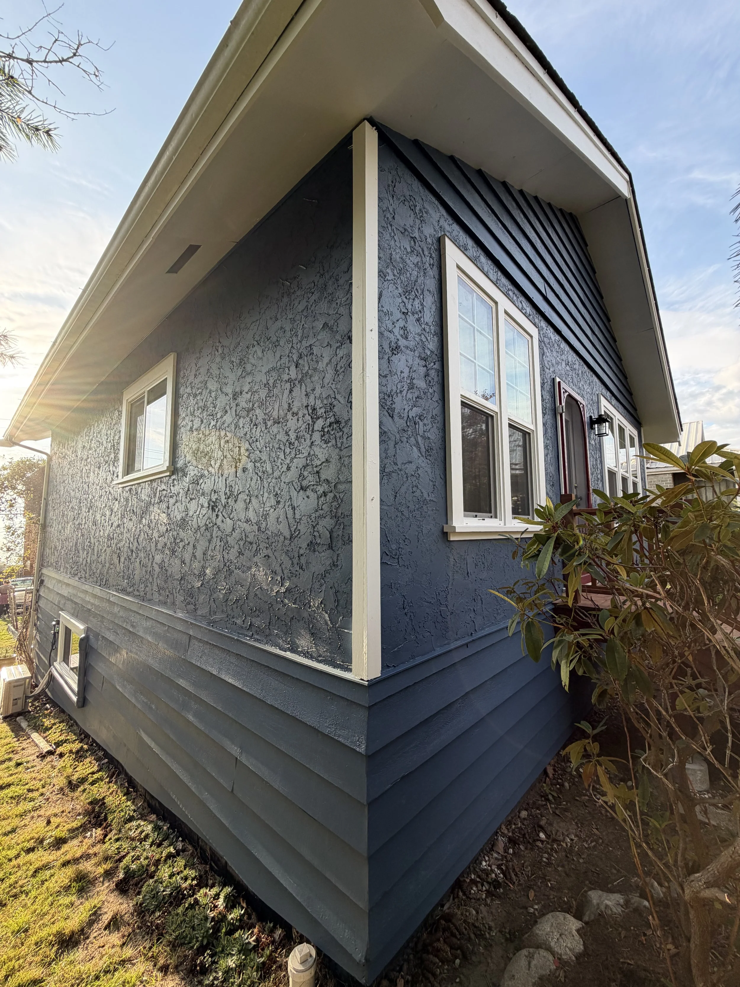 Close-up of the corner of a house with blue textured siding and white trim, showing three windows and a partially visible door, with a garden and sunlight in the background.