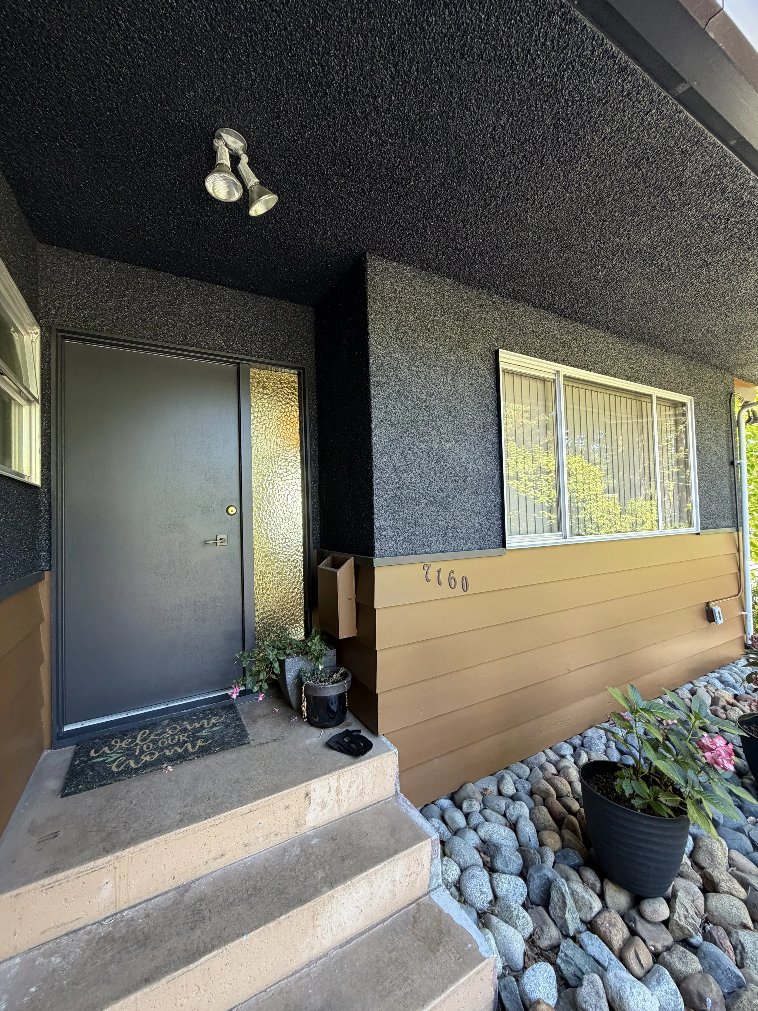The front porch of a house with a dark door, textured black walls, and a window. The house number is 9226. There are plants in pots and a doormat on the concrete step.