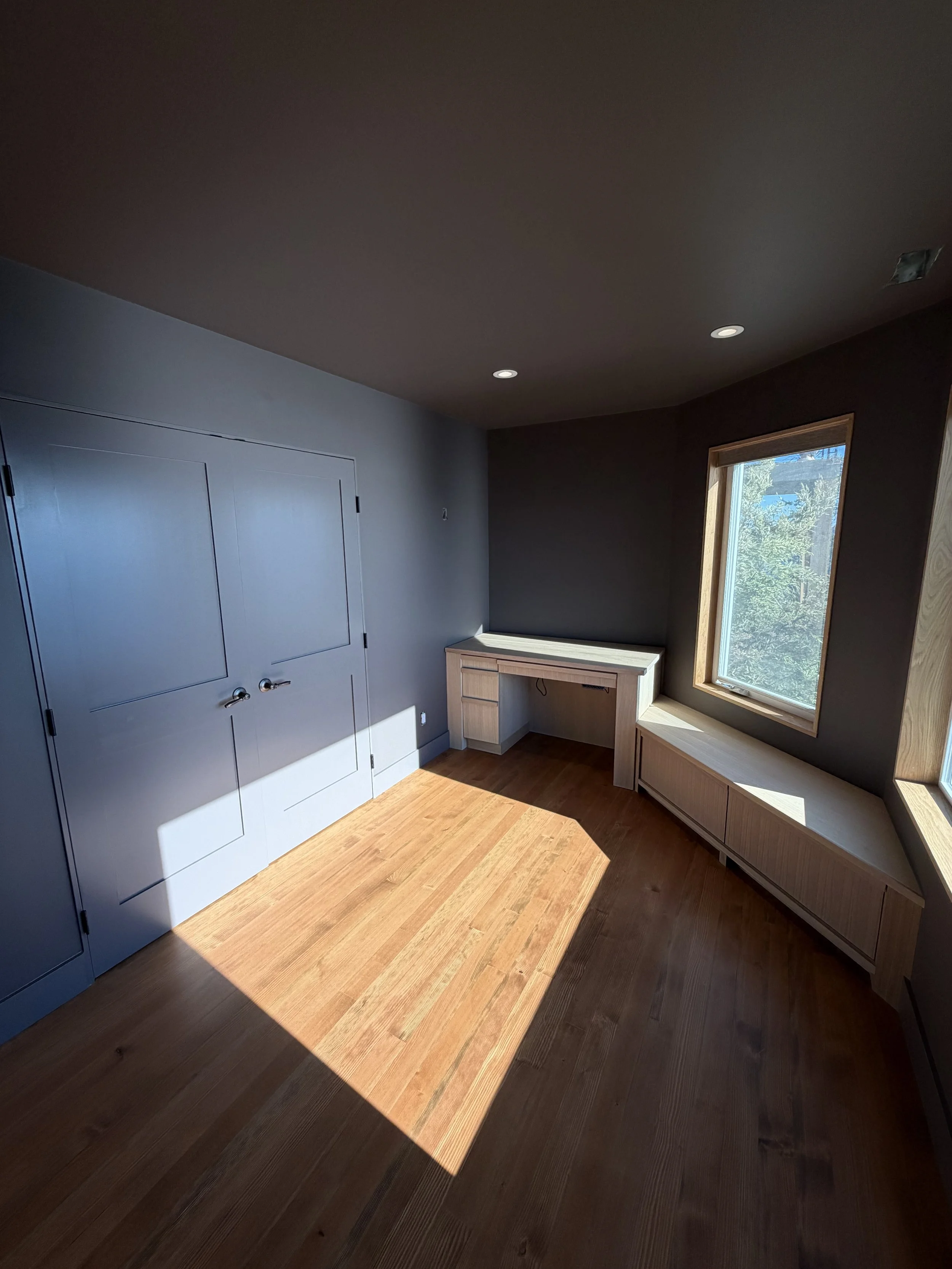A corner of a room with gray walls, a window with natural light, wooden floor, built-in wooden bench, and a small wooden desk next to the window.