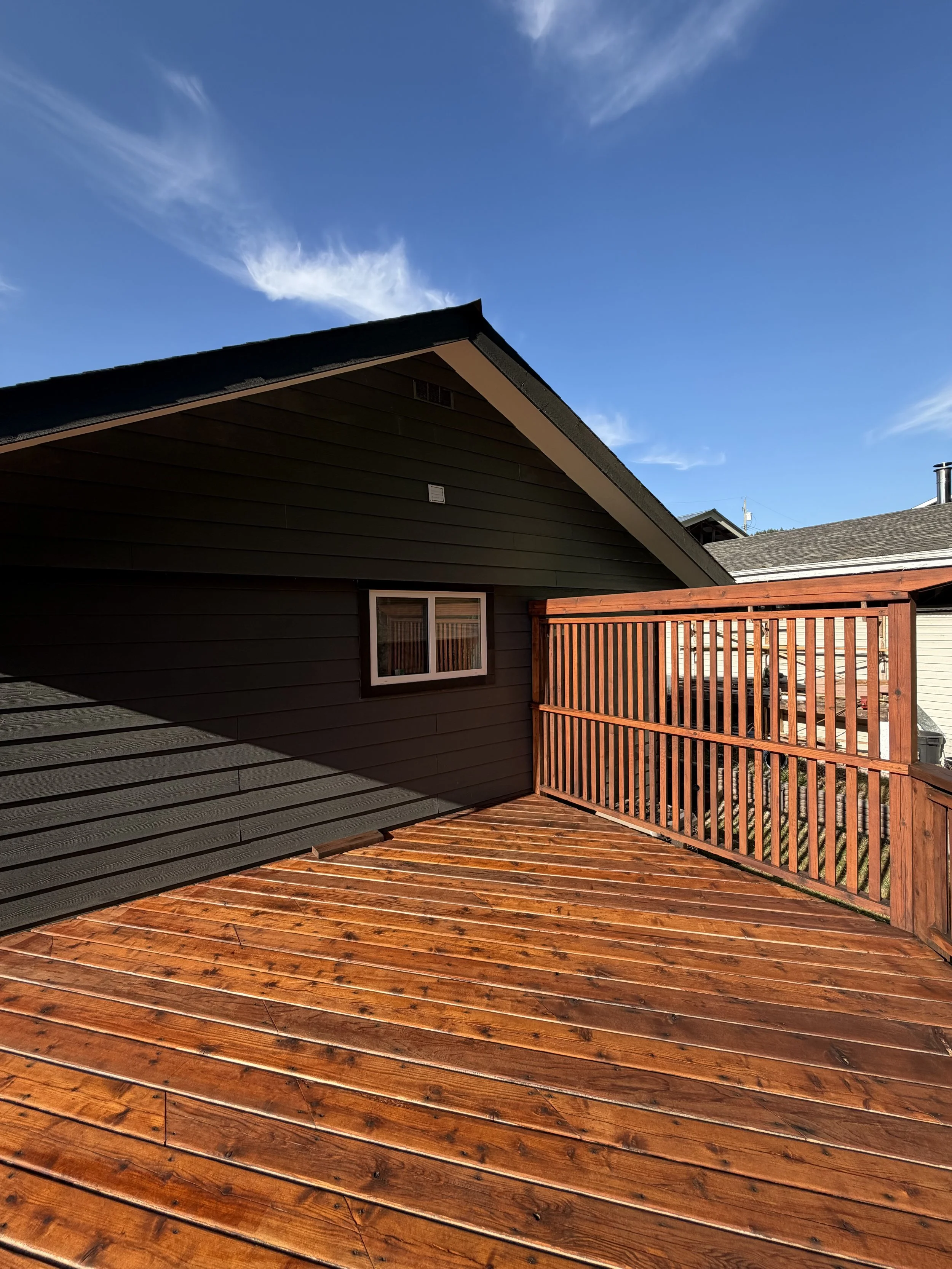 View of a wooden deck with a black house with a triangular roof and a window, a red wooden railing, and neighboring houses under a bright blue sky with some white clouds.