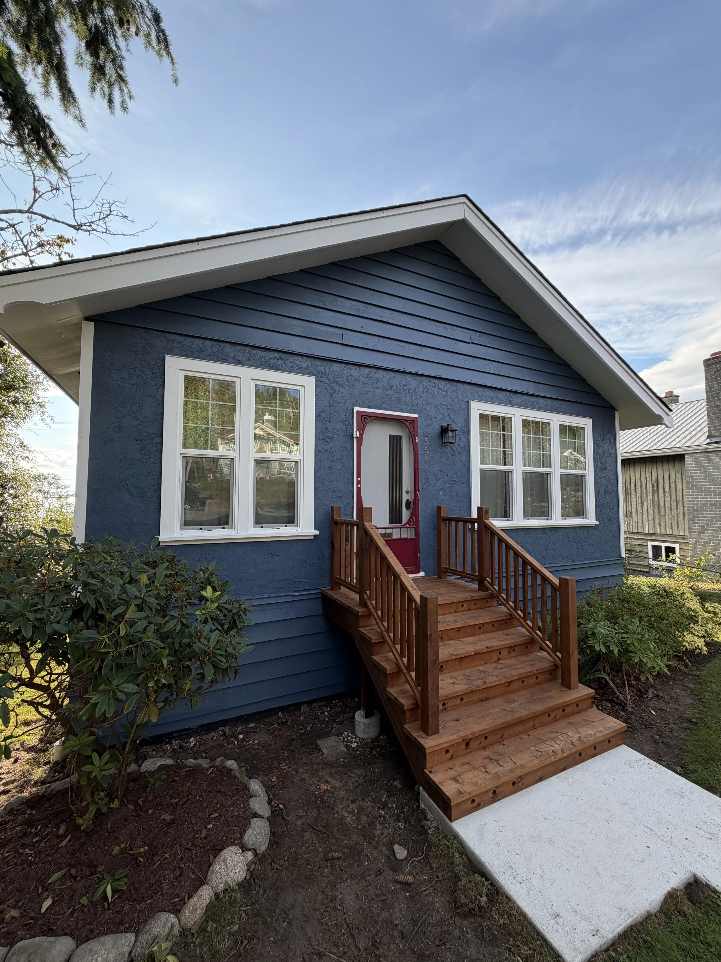 A house with blue exterior walls, white window frames, and a red front door with decorative trim. There is a small wooden staircase with railings leading up to the door. The house is surrounded by greenery and trees, with a partly cloudy sky overhead