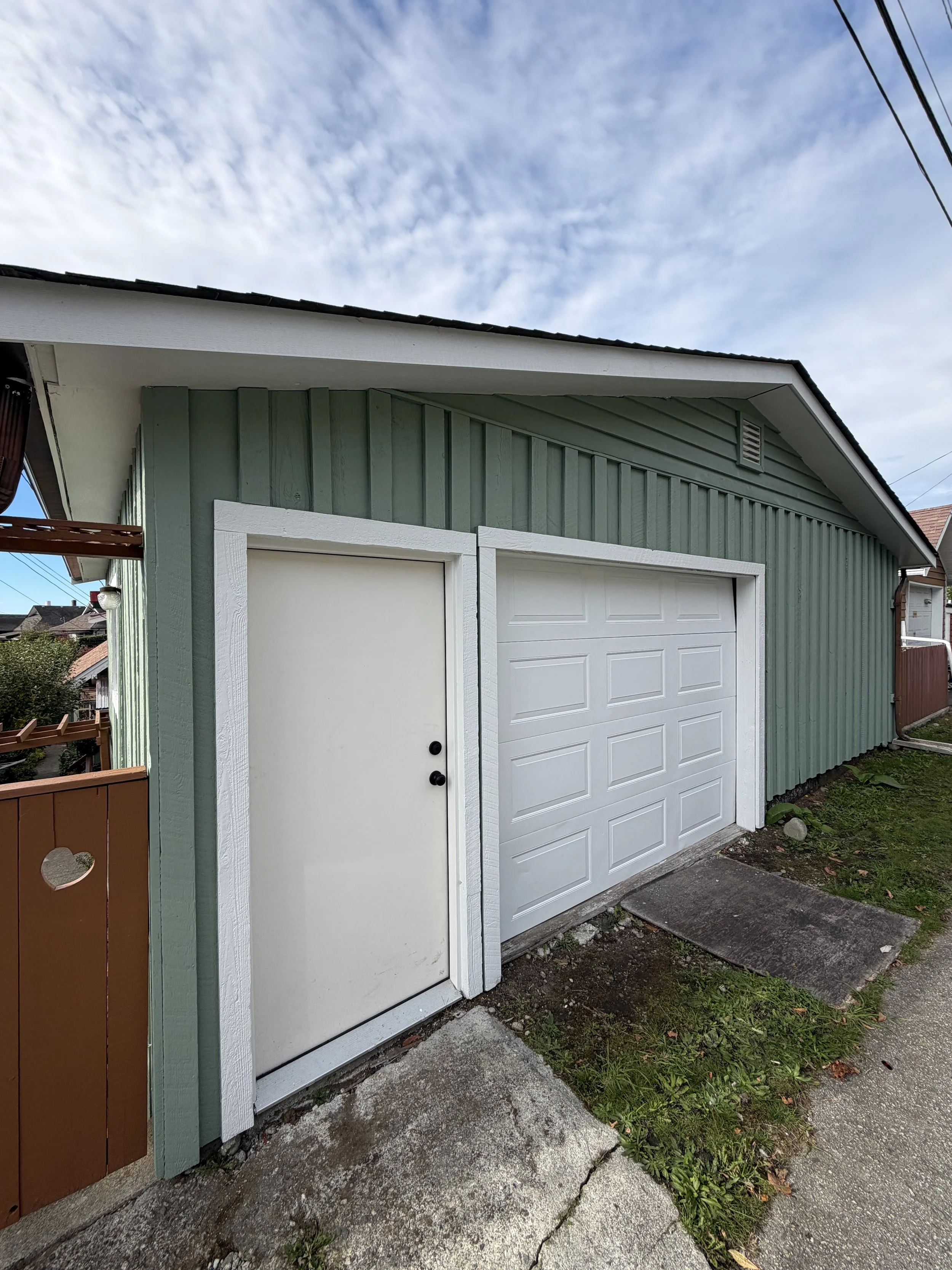 Exterior of a house with green vertical siding and white garage door, sidewalk in front, blue sky with clouds overhead.