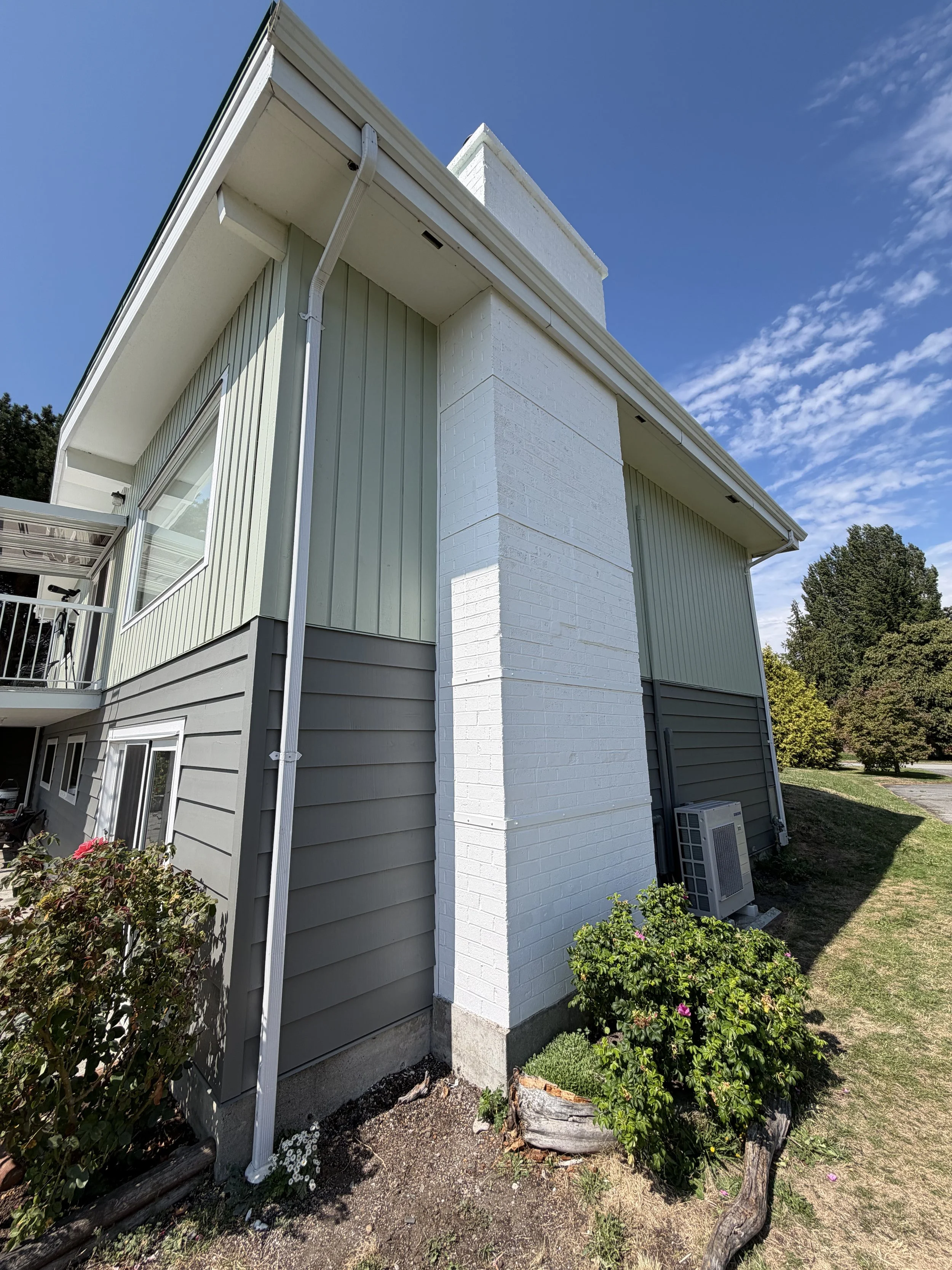 The side of a modern house with horizontal gray siding, white brick chimney, window, air conditioning unit, plants, and a small garden with flowers and a carved wooden post.