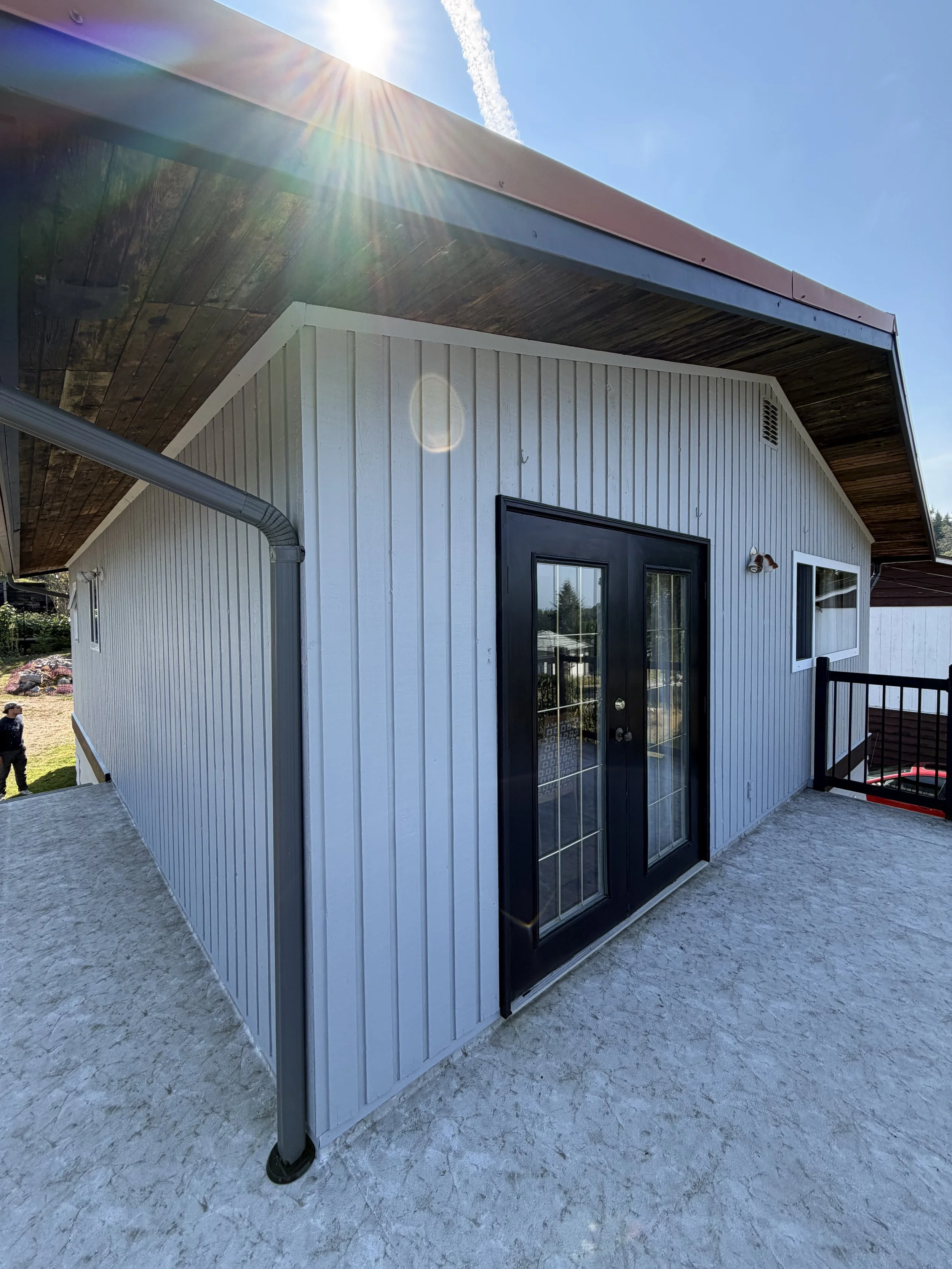 Exterior view of a modern house with white vertical siding, black framed glass door, and a small window, sun shining from above creating a lens flare, and a blue sky with a contrail in the background.