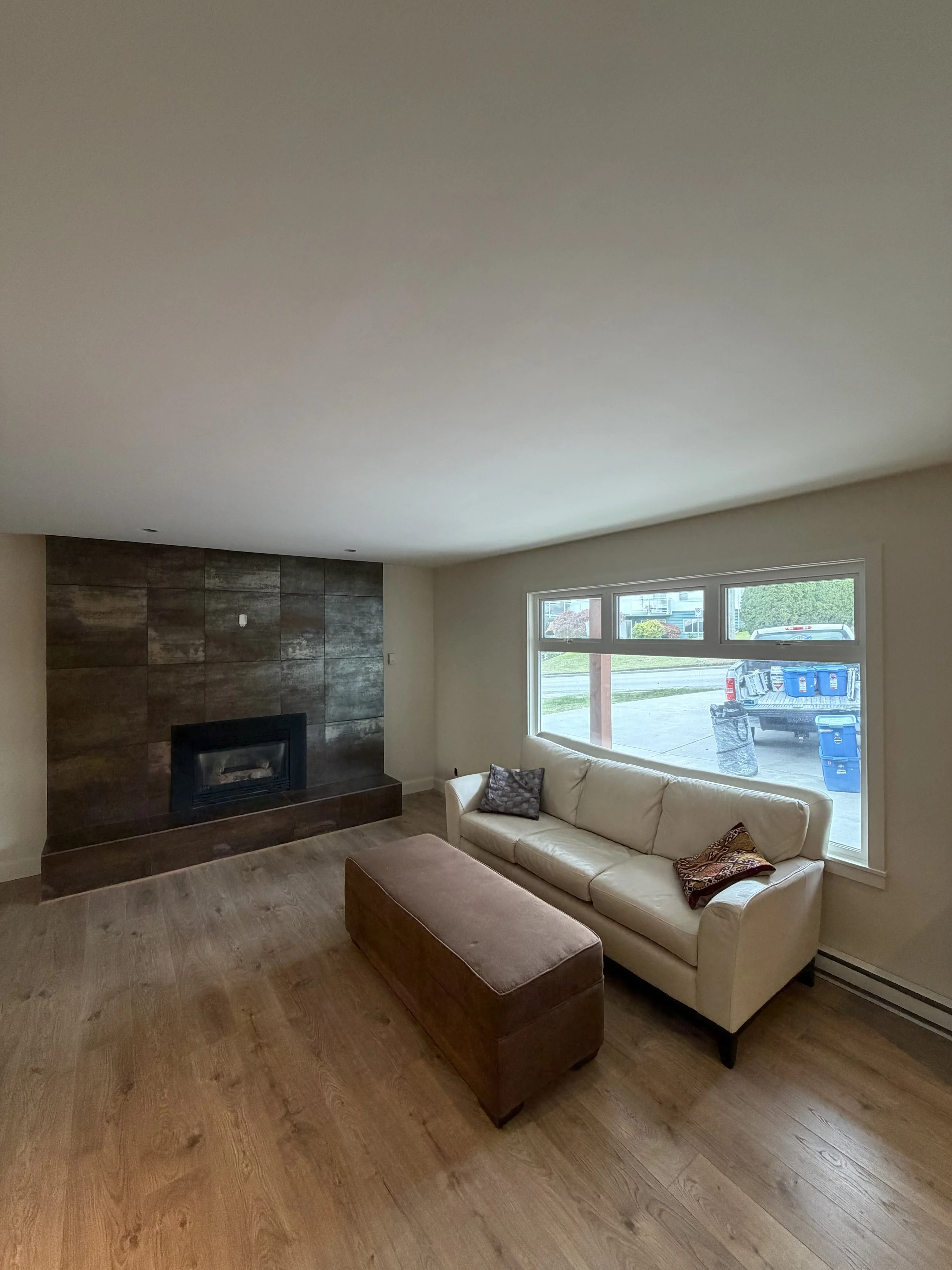 Living room with a white couch, a brown ottoman, wooden flooring, a wall with a wood-paneled fireplace, a window showing a truck and yard outside.
