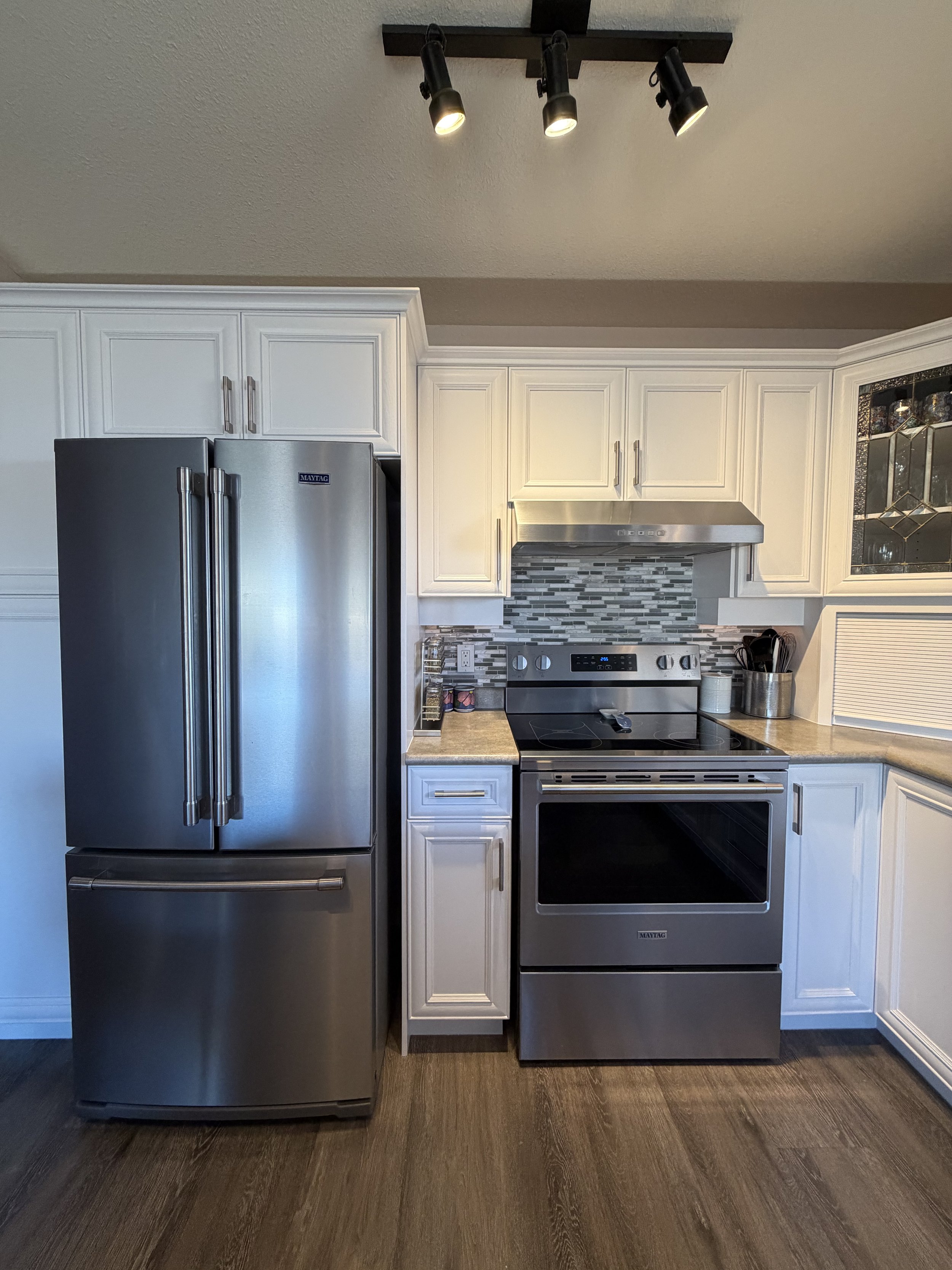 Modern kitchen with white cabinets, stainless steel Maytag refrigerator and electric range, mosaic tile backsplash, track lighting, and wood flooring
