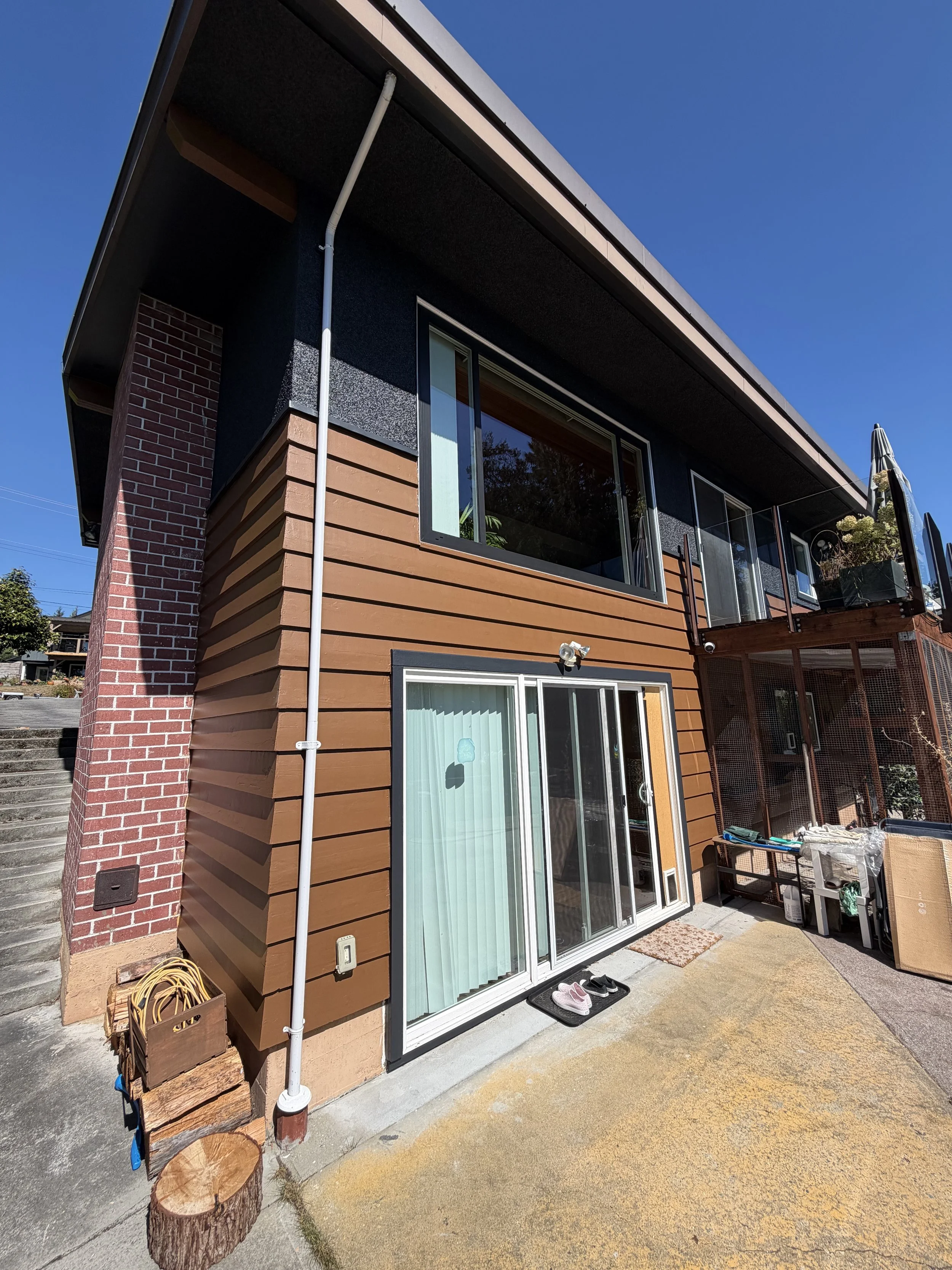 Side view of a modern two-story house with brown horizontal siding, brick accents, and black window frames. A sliding glass door opens to a small patio with shoes and outdoor furniture. A white downspout runs along the corner of the house, and a smal