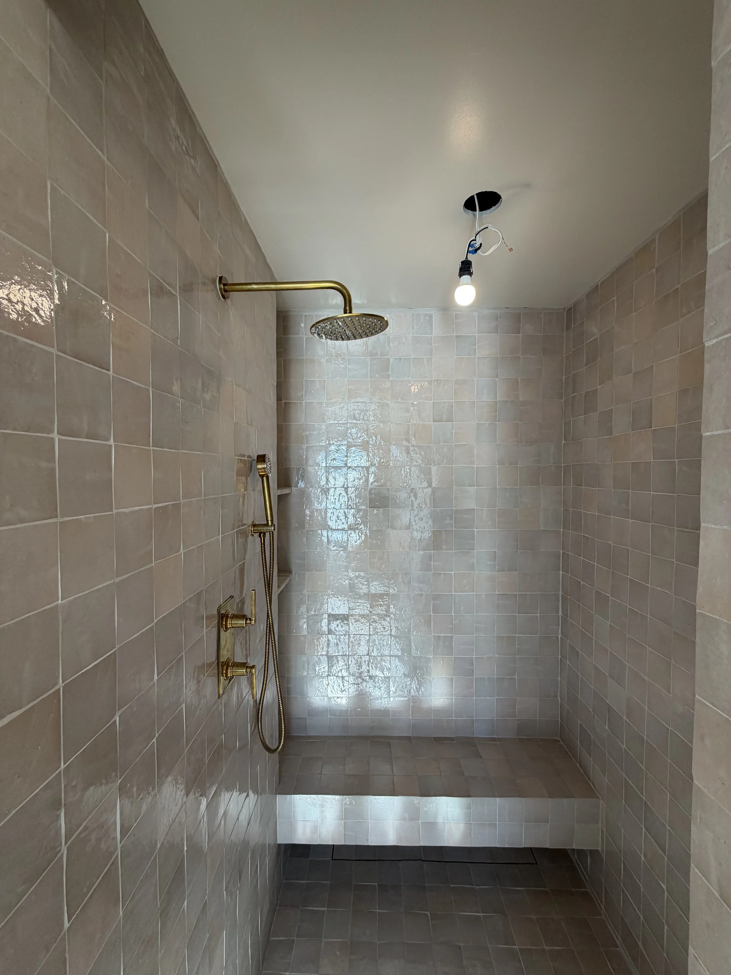 Empty shower area with beige tiles on the walls and floor, brass shower fixtures, and a ceiling light.