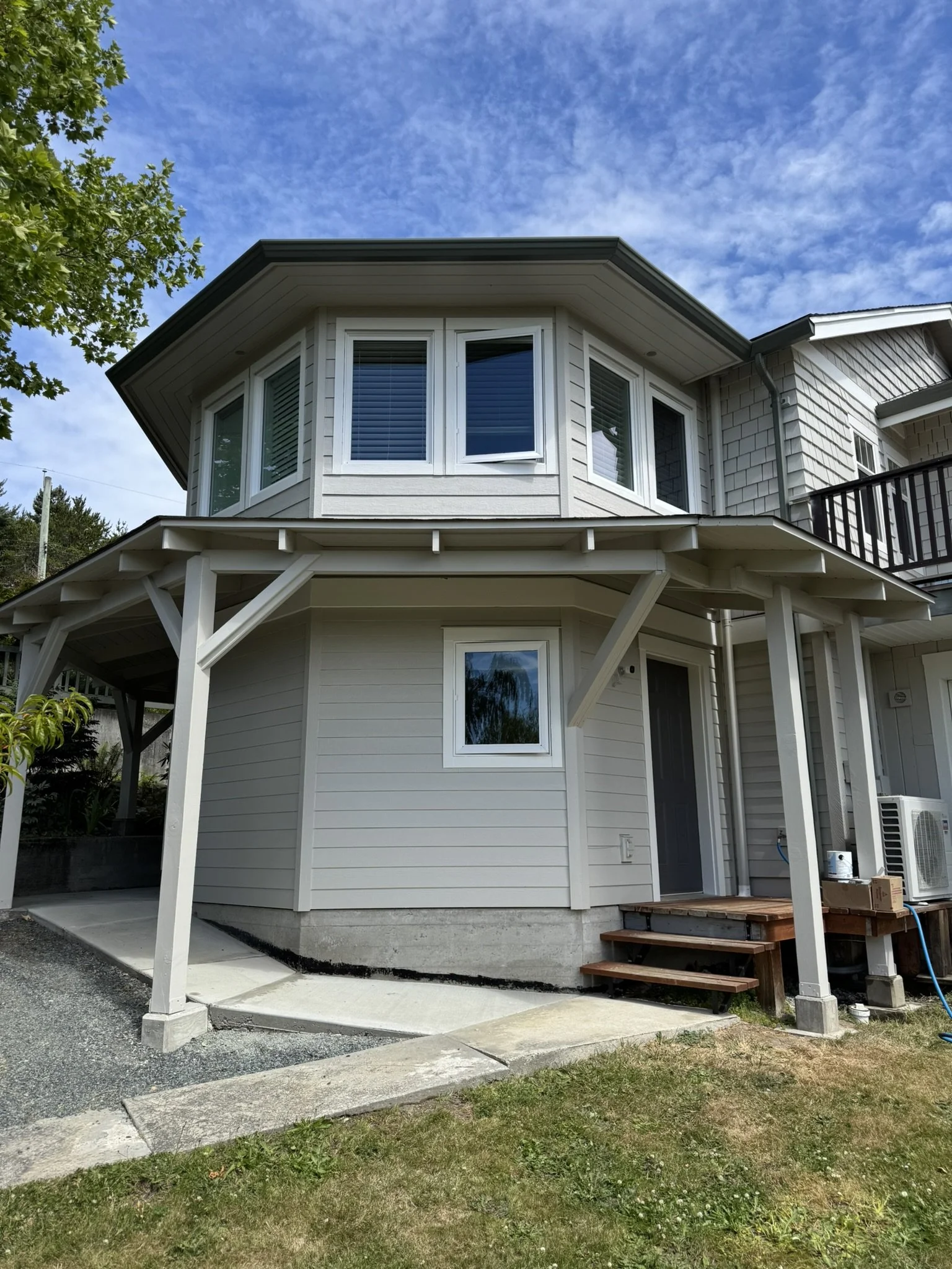 The image shows the exterior of a two-story house with white siding, with a small open porch supported by white posts. There is a window with blinds on the upper floor and a door on the lower floor. The area around the house has grass, gravel, and a 