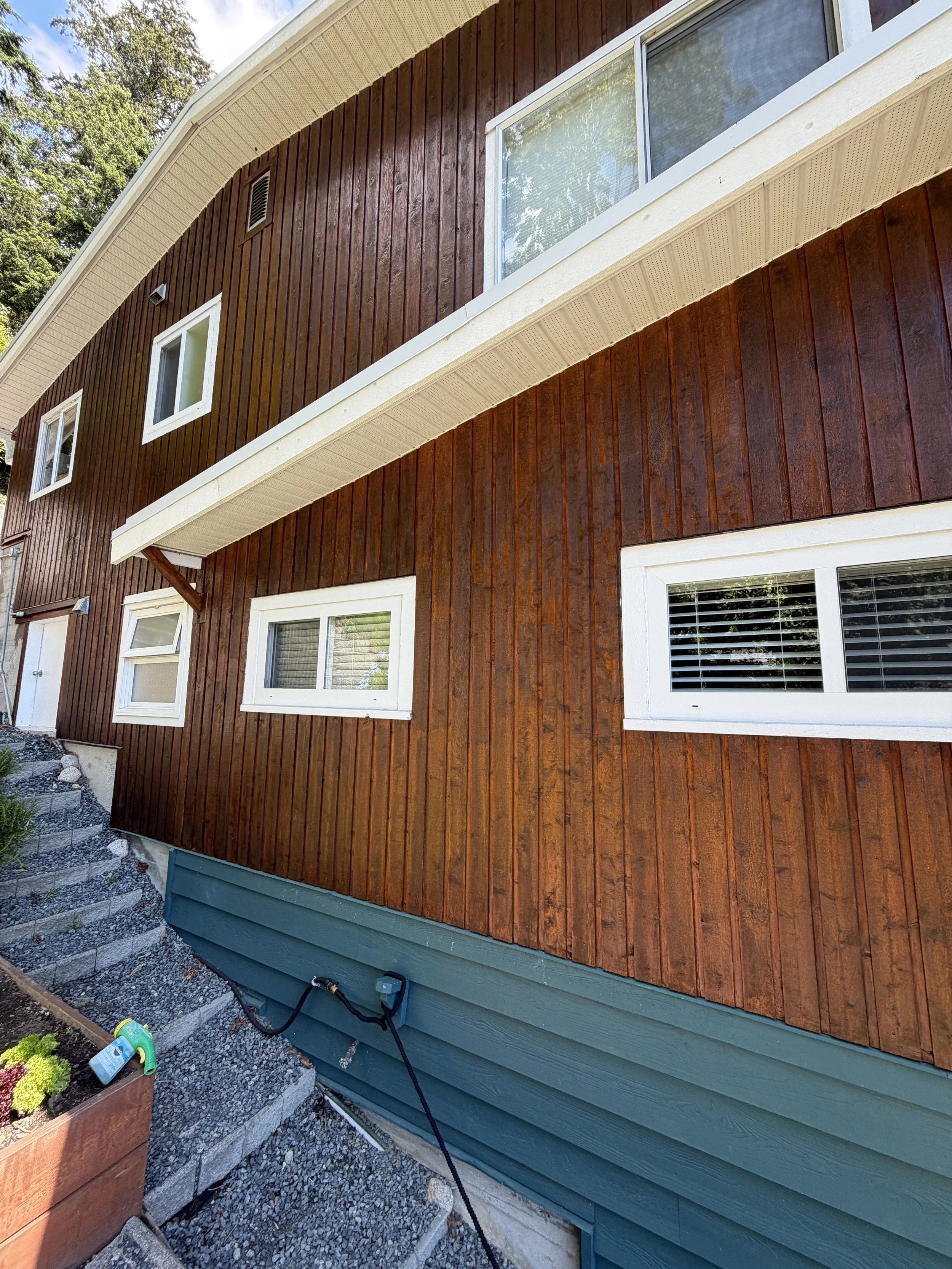 Side view of a house with brown wooden siding, white window frames, and a greenish-blue bottom section, with a flower bed and gardening supplies visible in the foreground.