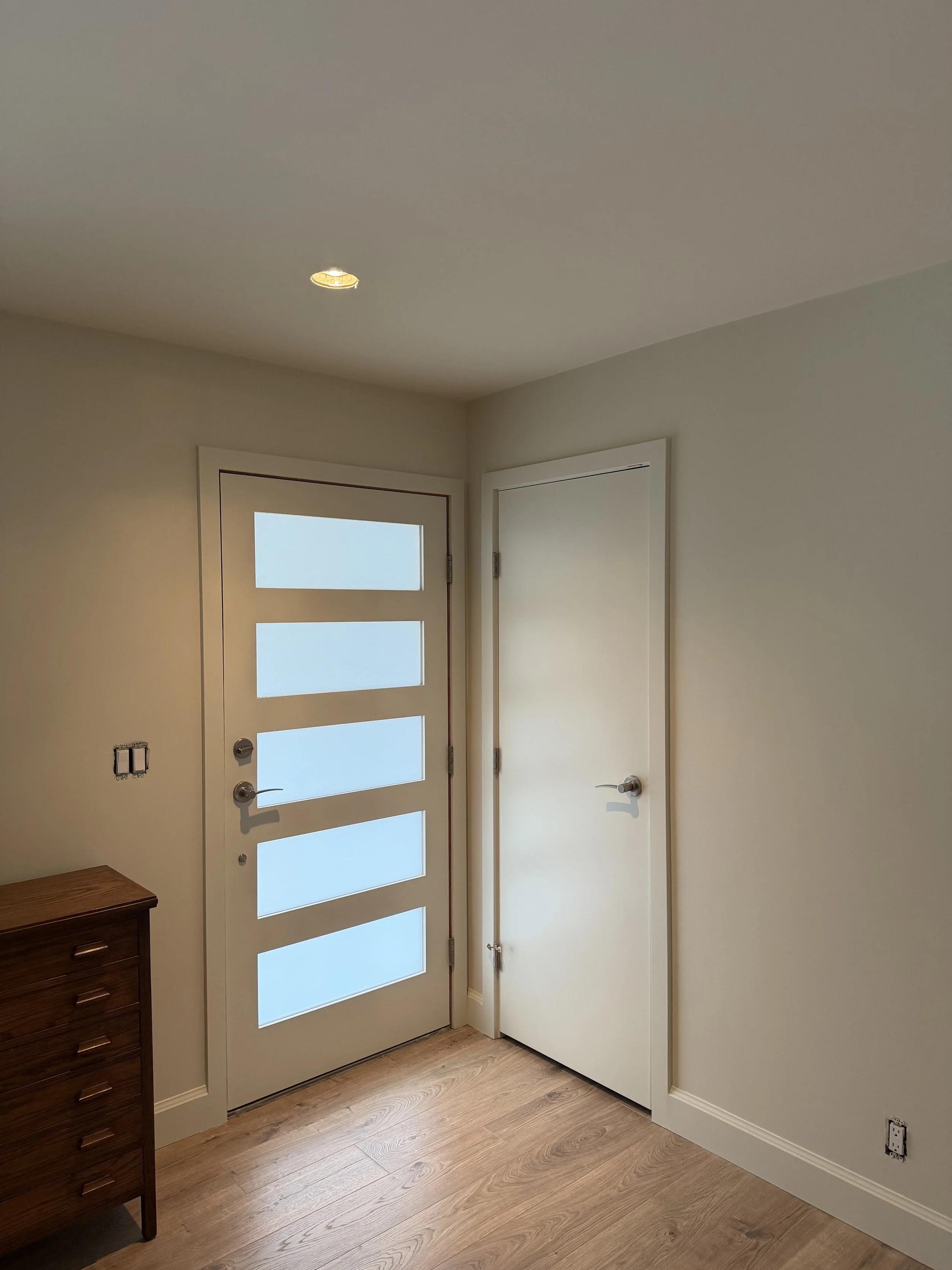 A room with a white door with frosted glass panels and a smaller white door, light wood flooring, and a dark wood chest of drawers.