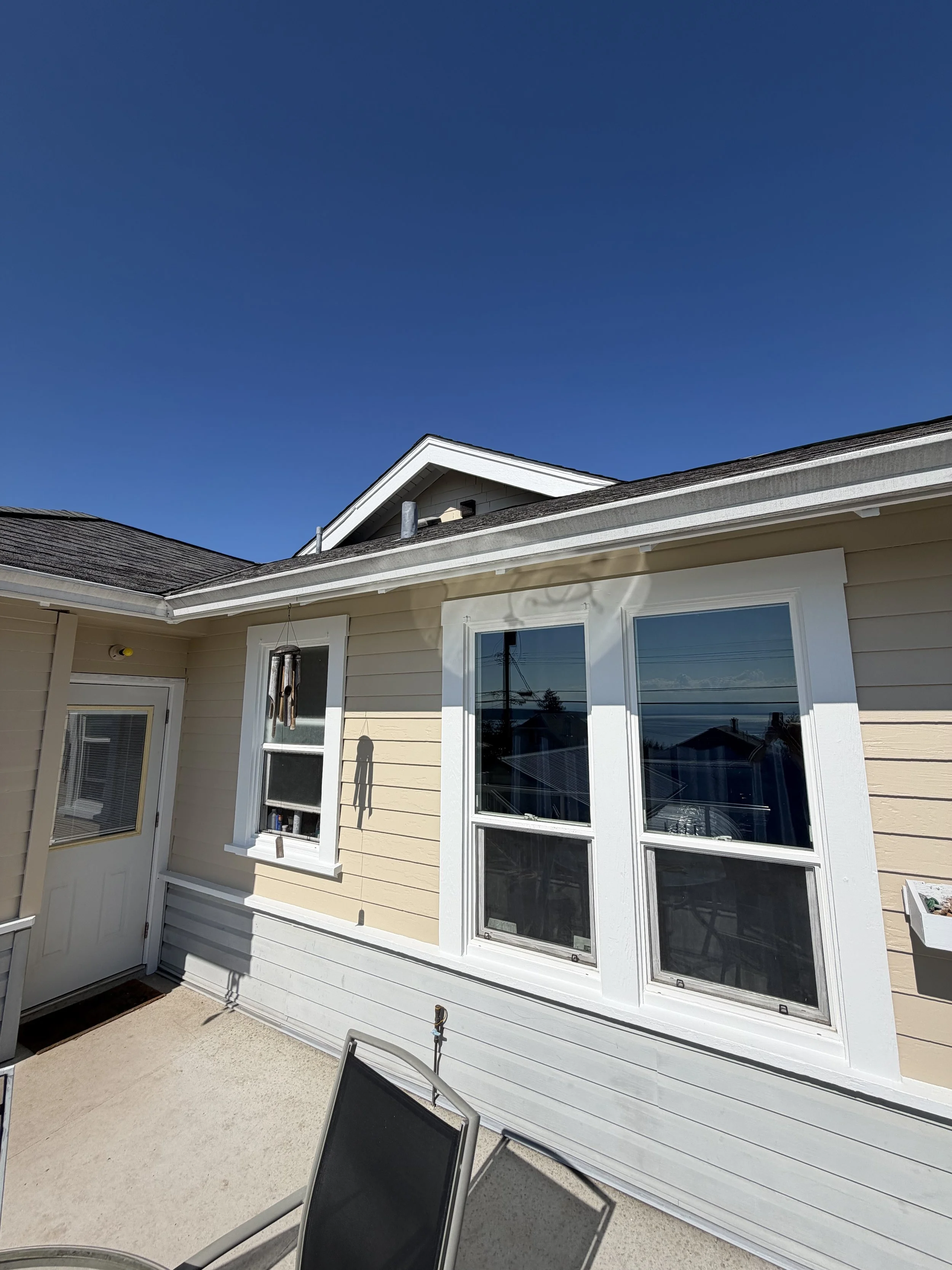 The image shows the exterior of a light yellow house with large windows and white trim. The house has a gabled roof. In the foreground, there is a concrete patio with two black outdoor chairs and a small metal structure, possibly a lantern or pest co