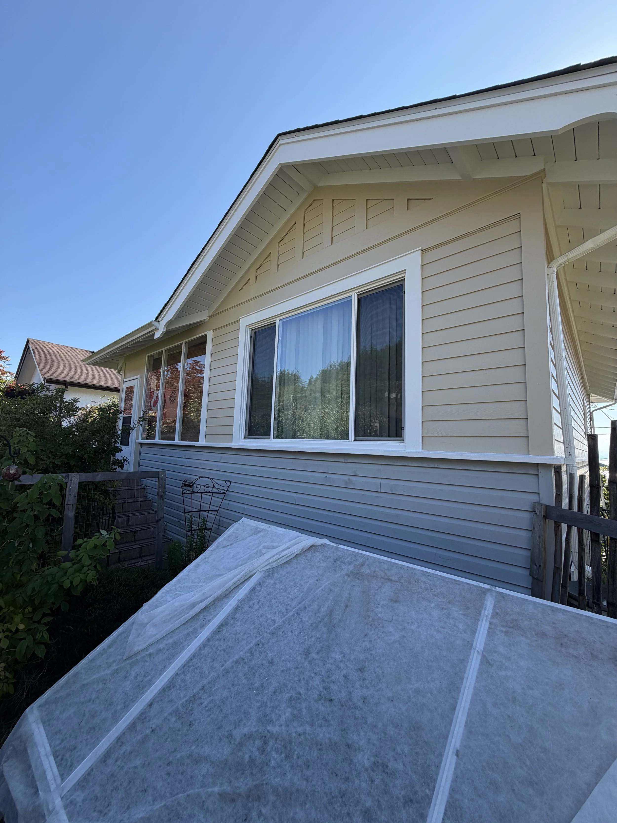The image shows the side of a beige house with large sliding windows and a white trim. There is a small garden and a gray cover in the foreground, with a clear blue sky above.