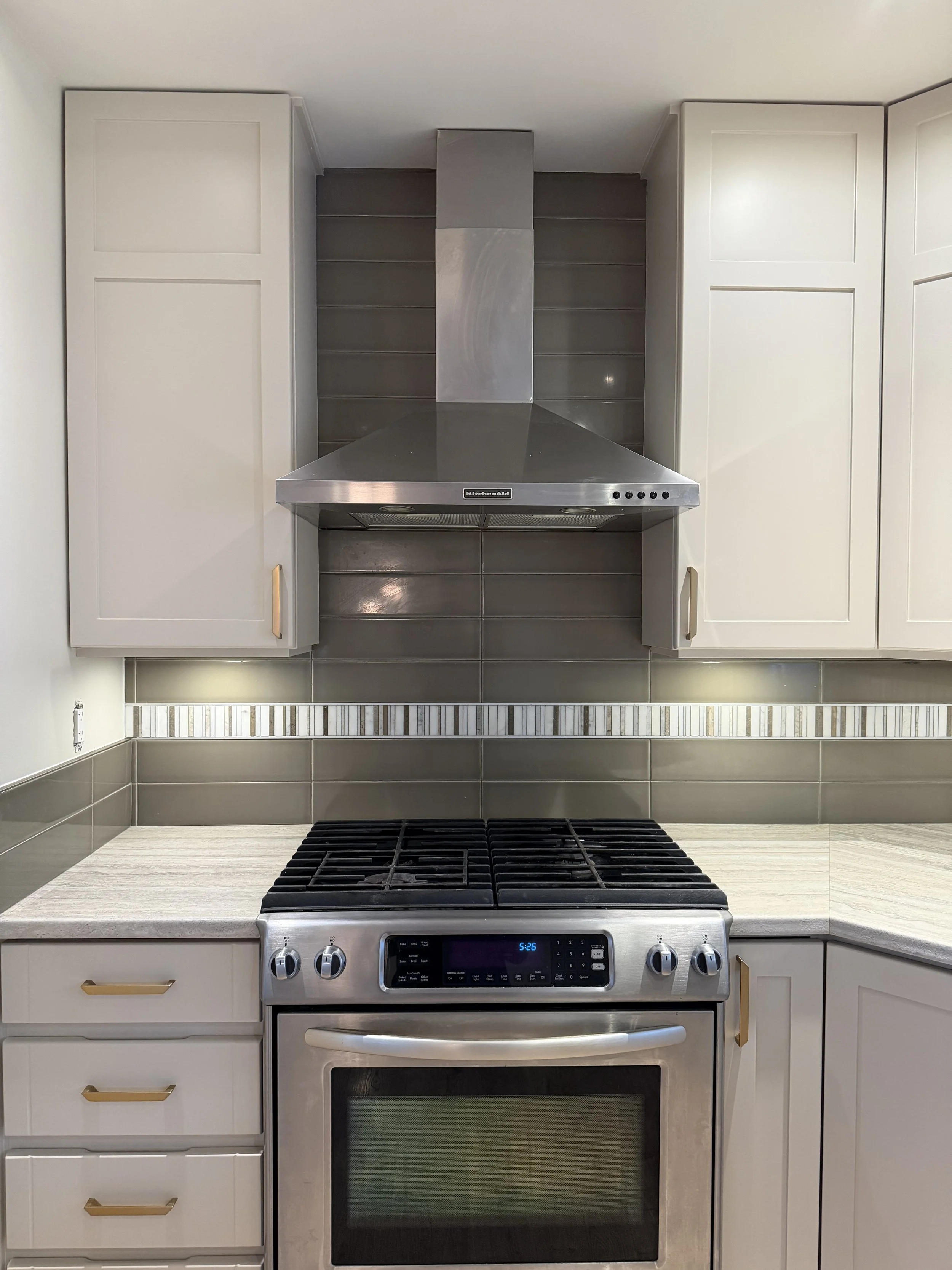 Modern kitchen stove with stainless steel oven, black stovetop, white cabinets with gold handles, gray tiled backsplash, and countertop.