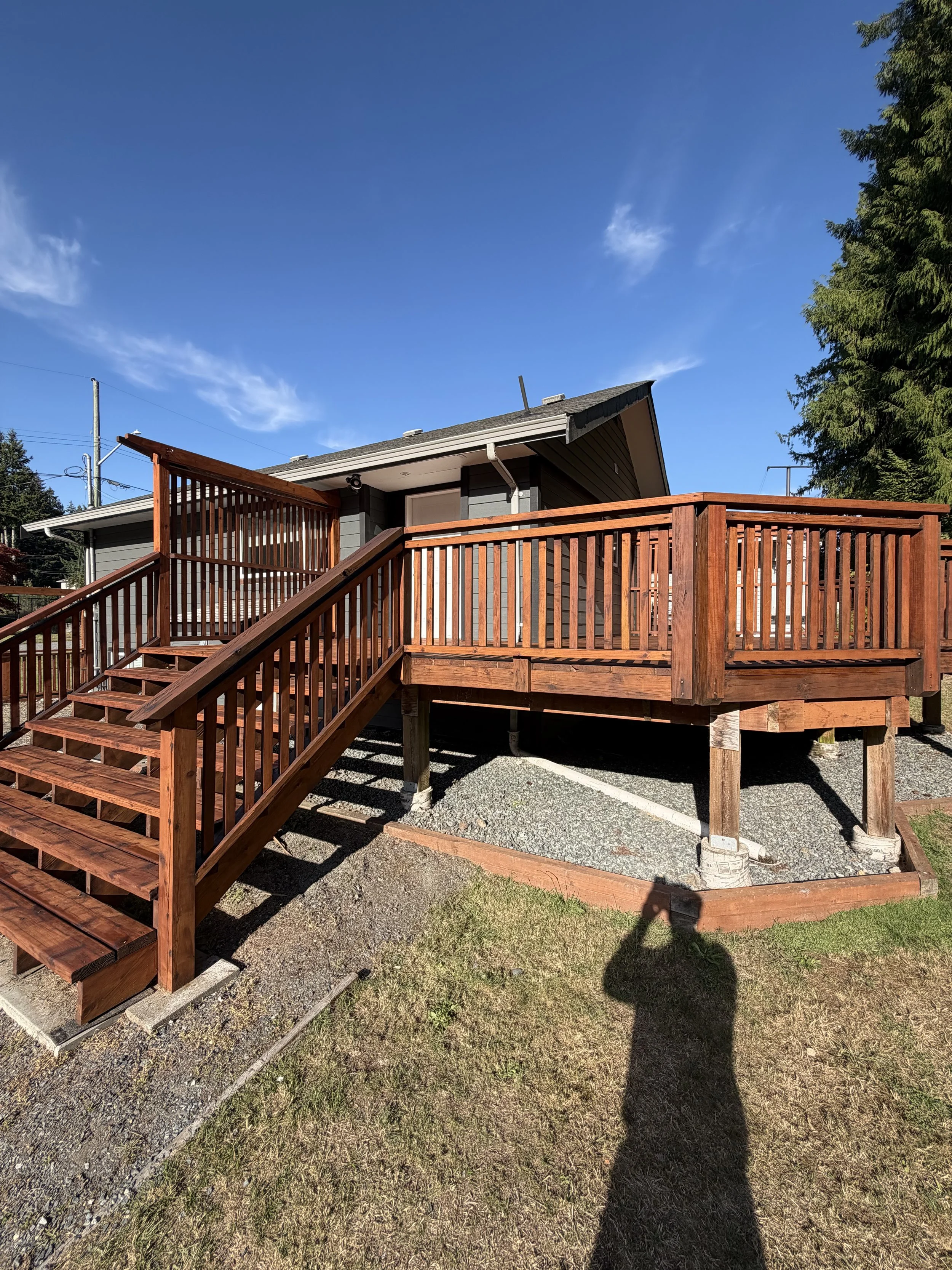A house with a wooden deck and stairs, under a blue sky with a few clouds, surrounded by green trees and grass.