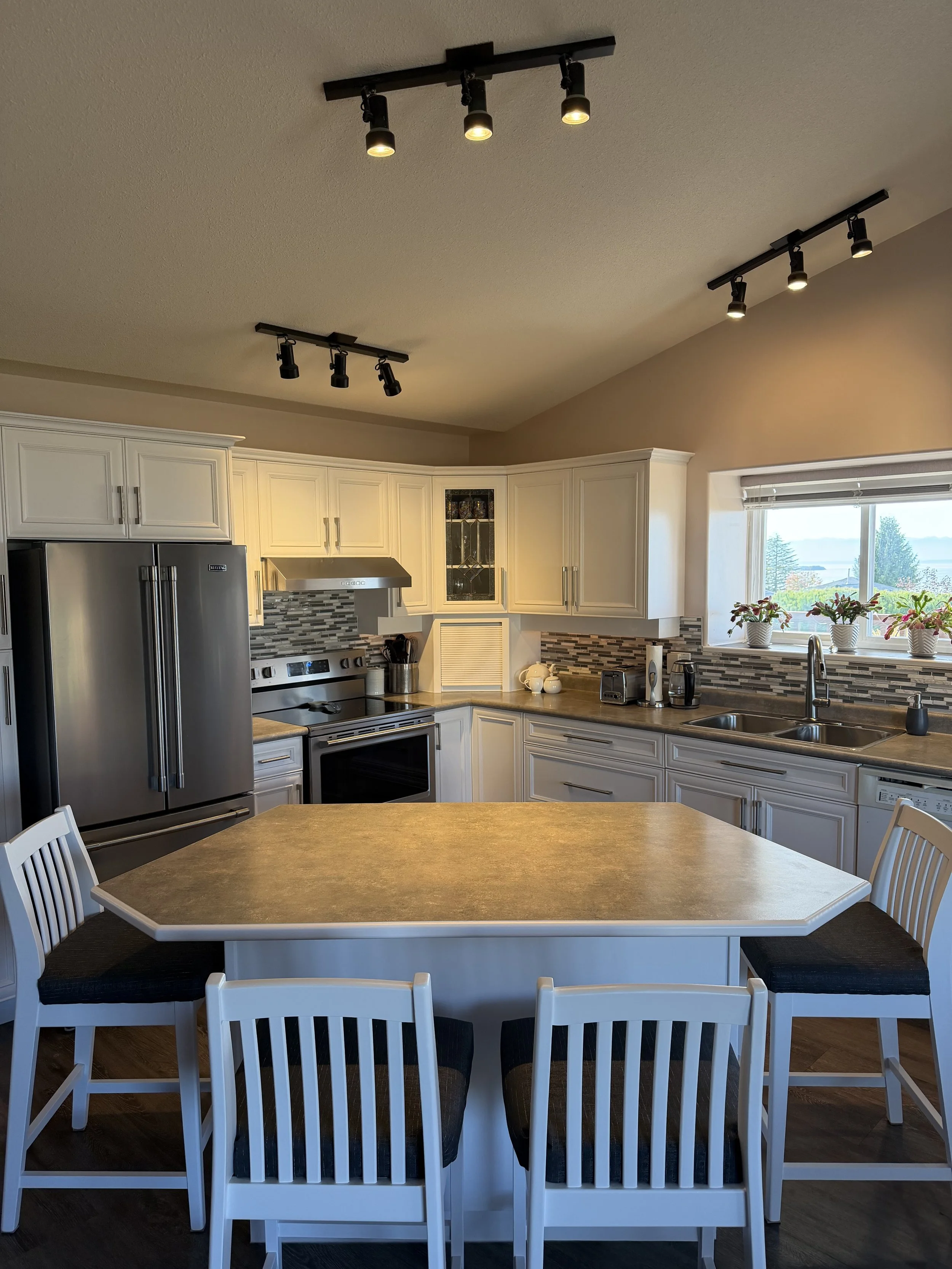 Bright kitchen with white cabinets, stainless steel appliances, track lighting, a large dining island, and a window overlooking the outdoors.