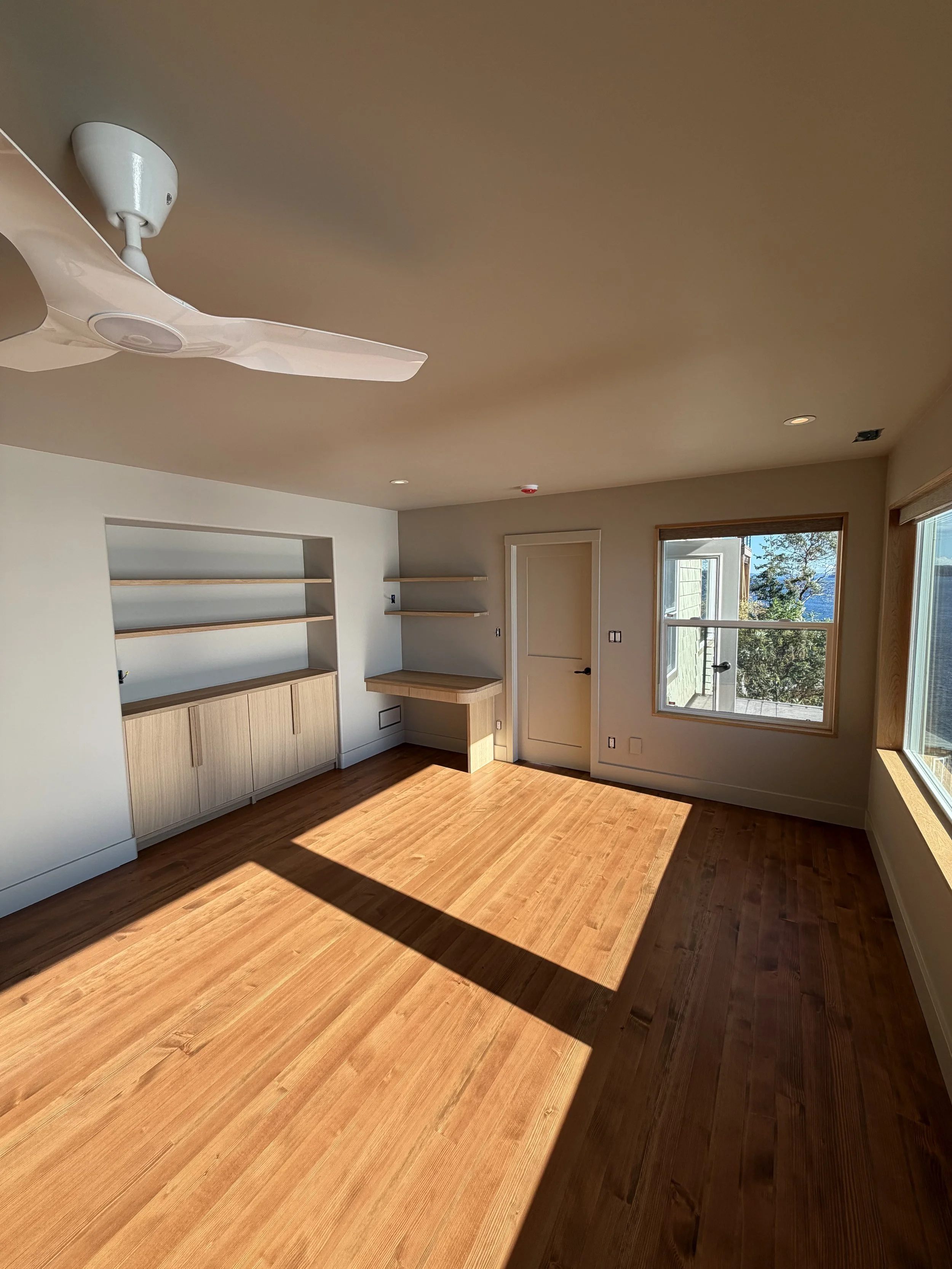 Empty room with wooden flooring, large window, built-in shelves, and a ceiling fan.
