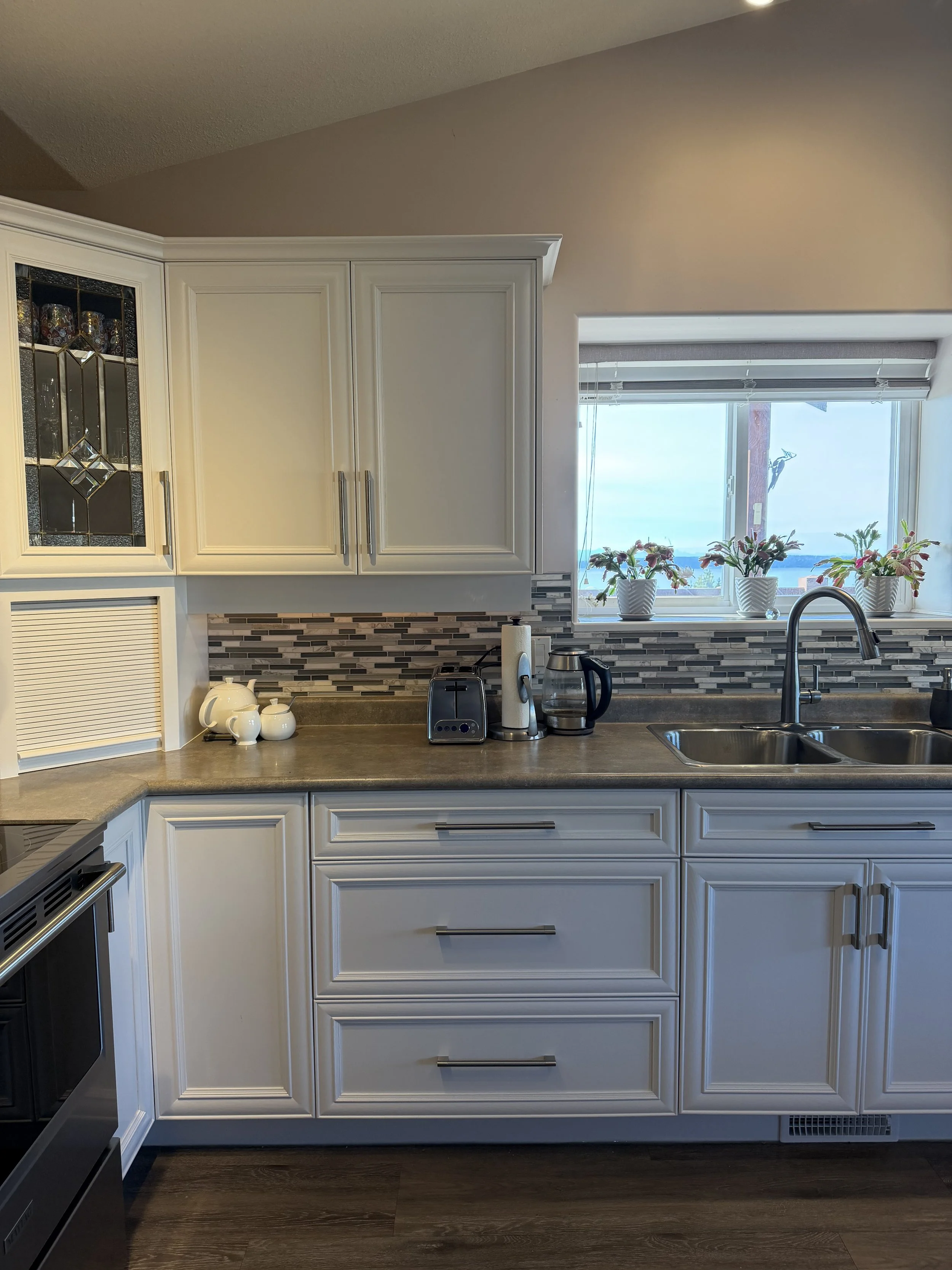 Bright kitchen with white cabinets, mosaic backsplash, stainless sink, countertop appliances, potted plants, and a window overlooking a lake.