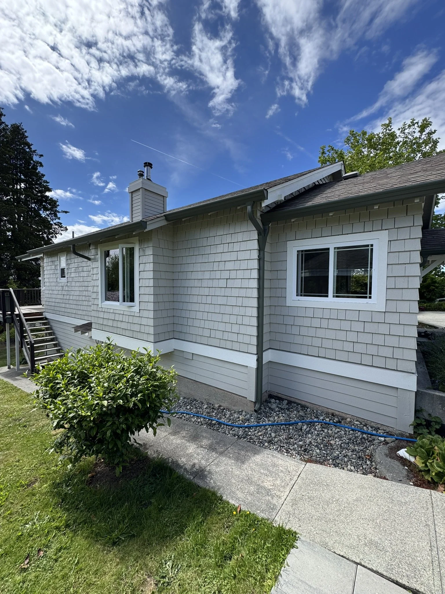Back view of a house with white siding, two windows, a chimney, a stairway on the left, and a garden with green bushes. There is a concrete walkway and a patch of grass. The sky is blue with scattered clouds and a faint contrail.