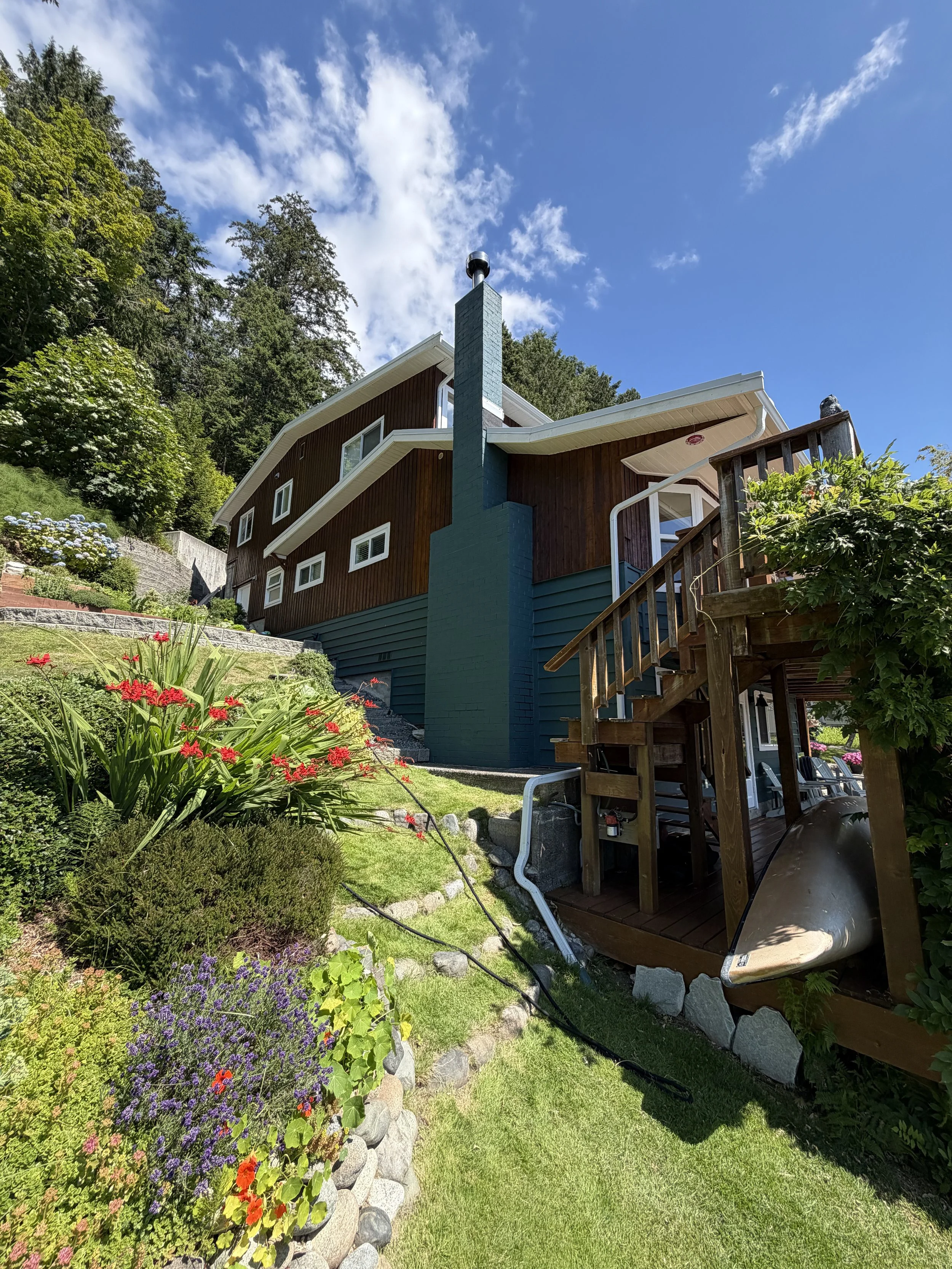 A backyard garden with a house having a sloped roof, a chimney, and a wooden deck. There are colorful flowers and green plants, with a kayak resting underneath the deck. The sky is bright with some clouds.