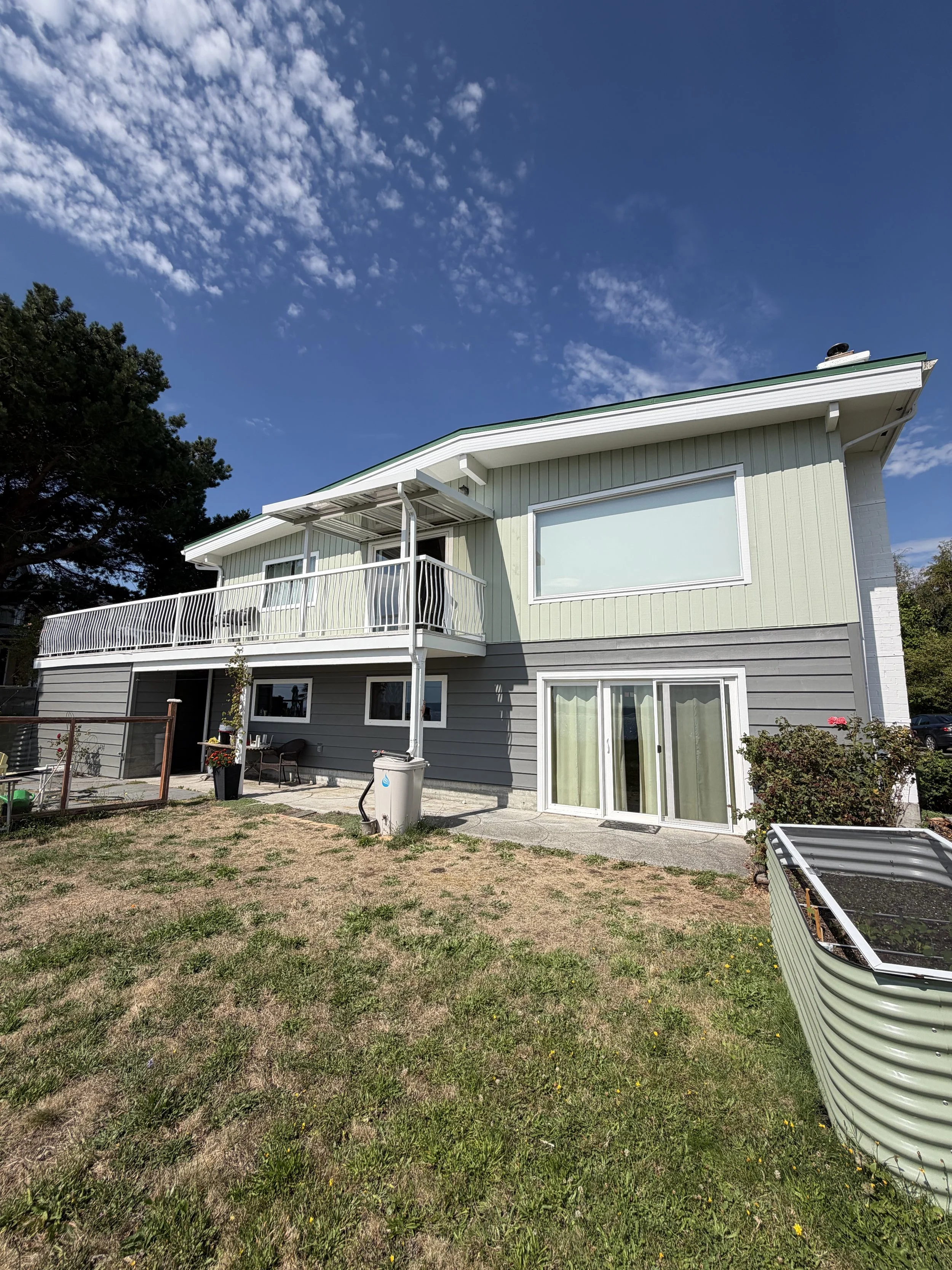 A two-story house with light green and gray siding, a balcony with white railing, large windows, and a patio area with some furniture and a small lawn with patches of grass and dirt, under a blue sky with clouds.