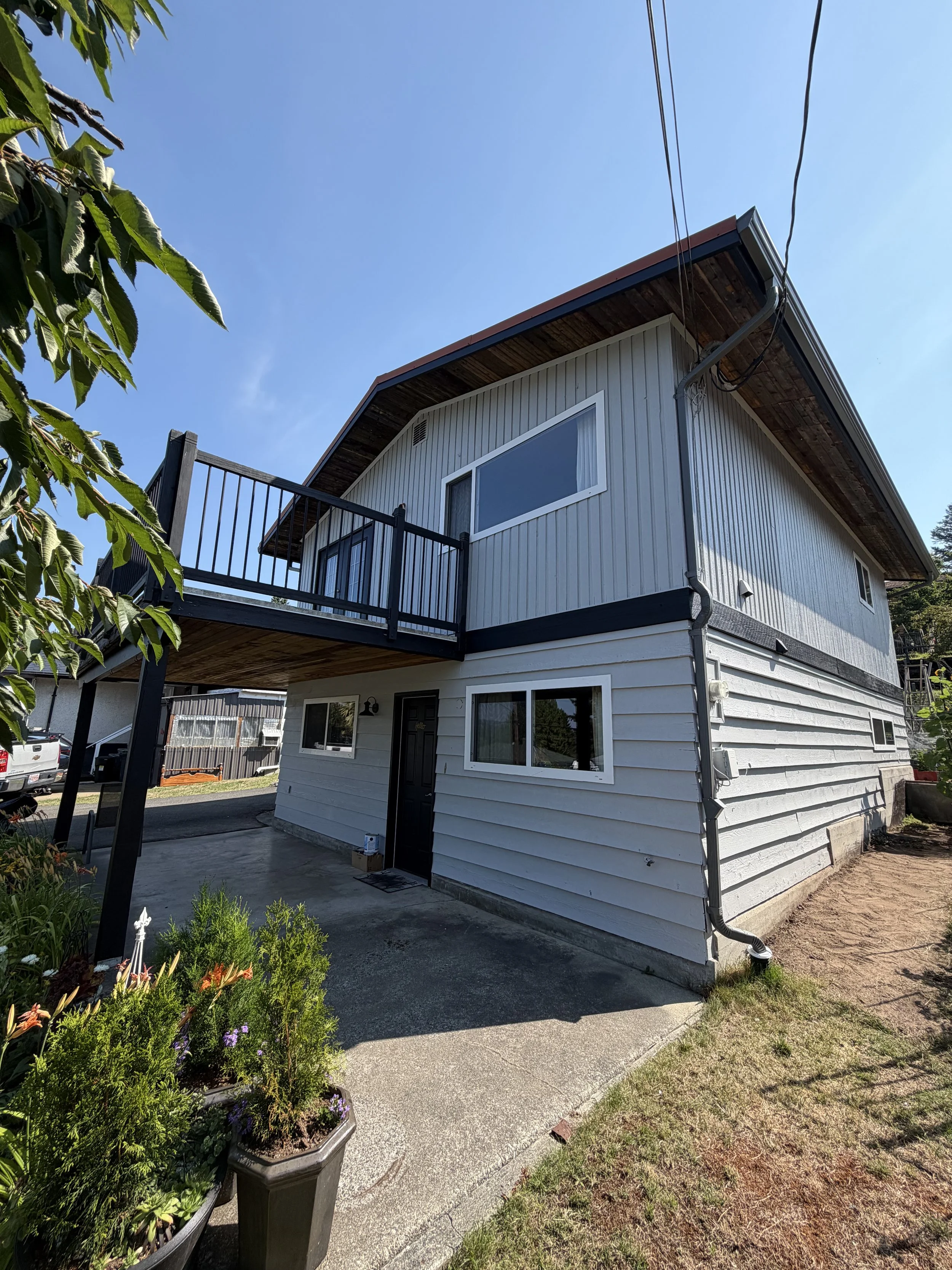 A two-story house with a white exterior, black accents, and a small balcony on the upper level. There are green plants in pots in the foreground, and a concrete walkway leading to the front door. The sky is clear and blue.
