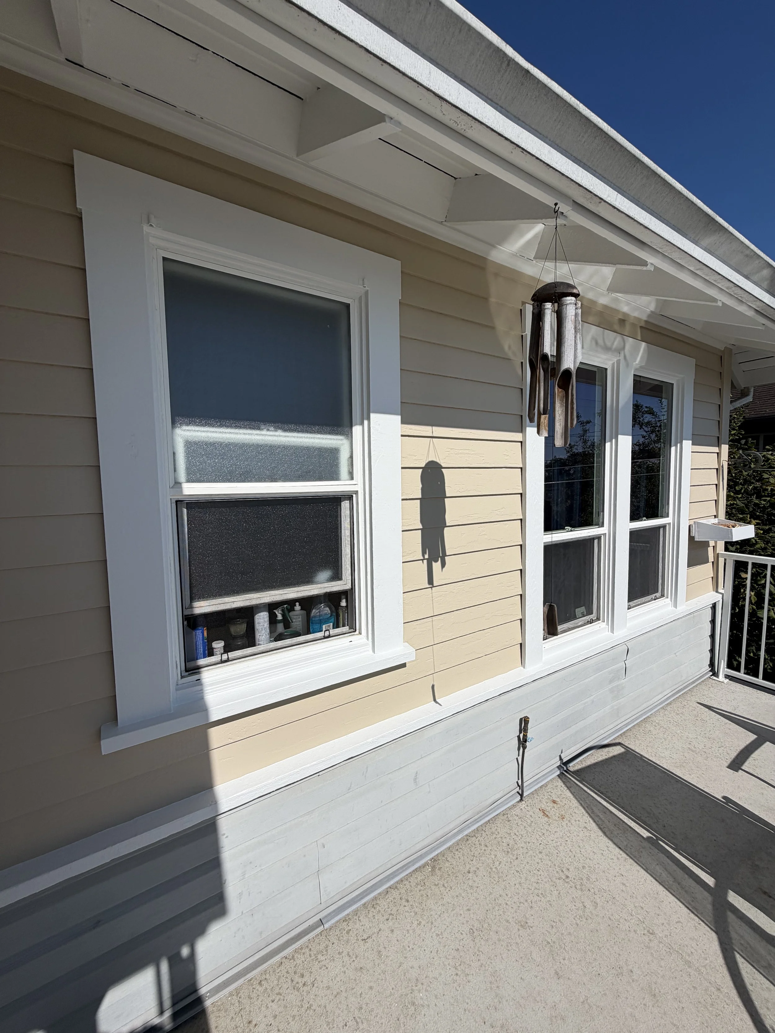 The image shows the exterior of a house with beige horizontal siding and white trim around two windows. A wind chime hangs near the windows, and there's a small section of a railing and door frame on the right. The sky is clear and blue.