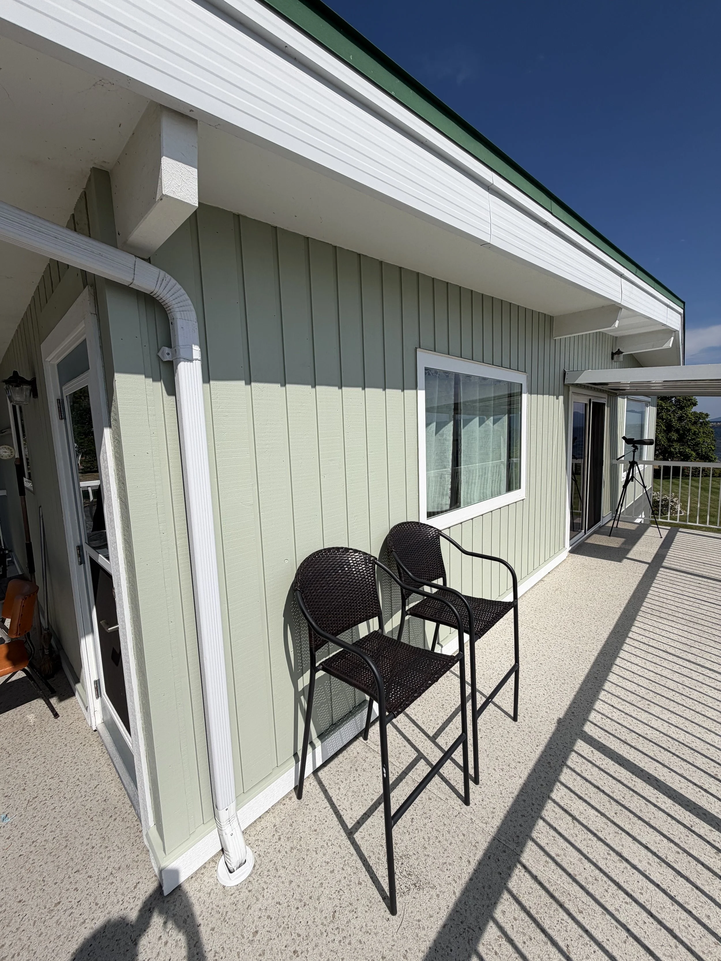 A balcony with three black outdoor chairs, a sliding glass door, and a railing overlooking trees and water in the background. The exterior wall is painted light green with vertical siding, and there are white window and door trims. The ceiling overha