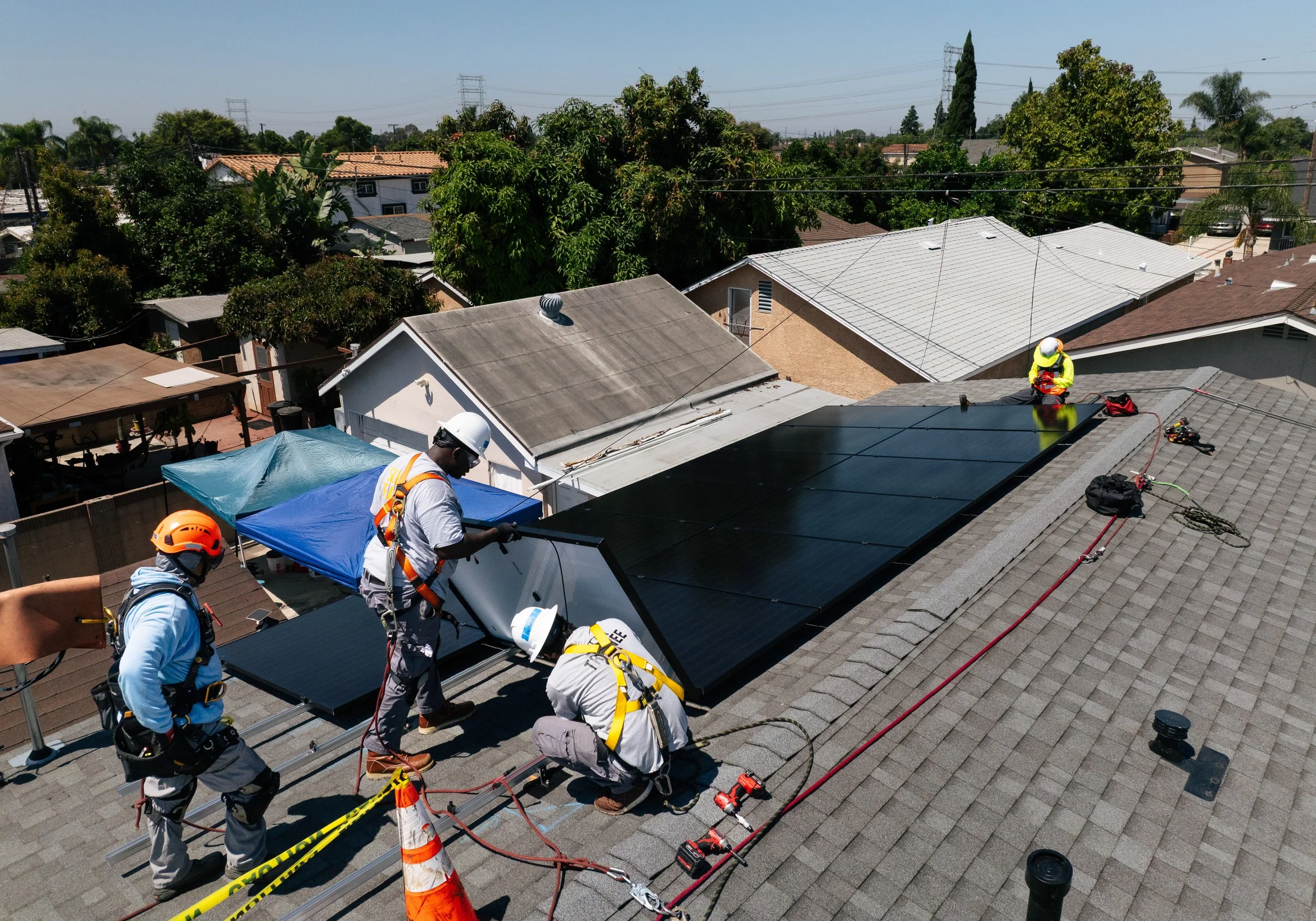 GRID Alternatives installers secure solar panels on a residential rooftop in Los Angeles, California, with the surrounding neighborhood visible below.