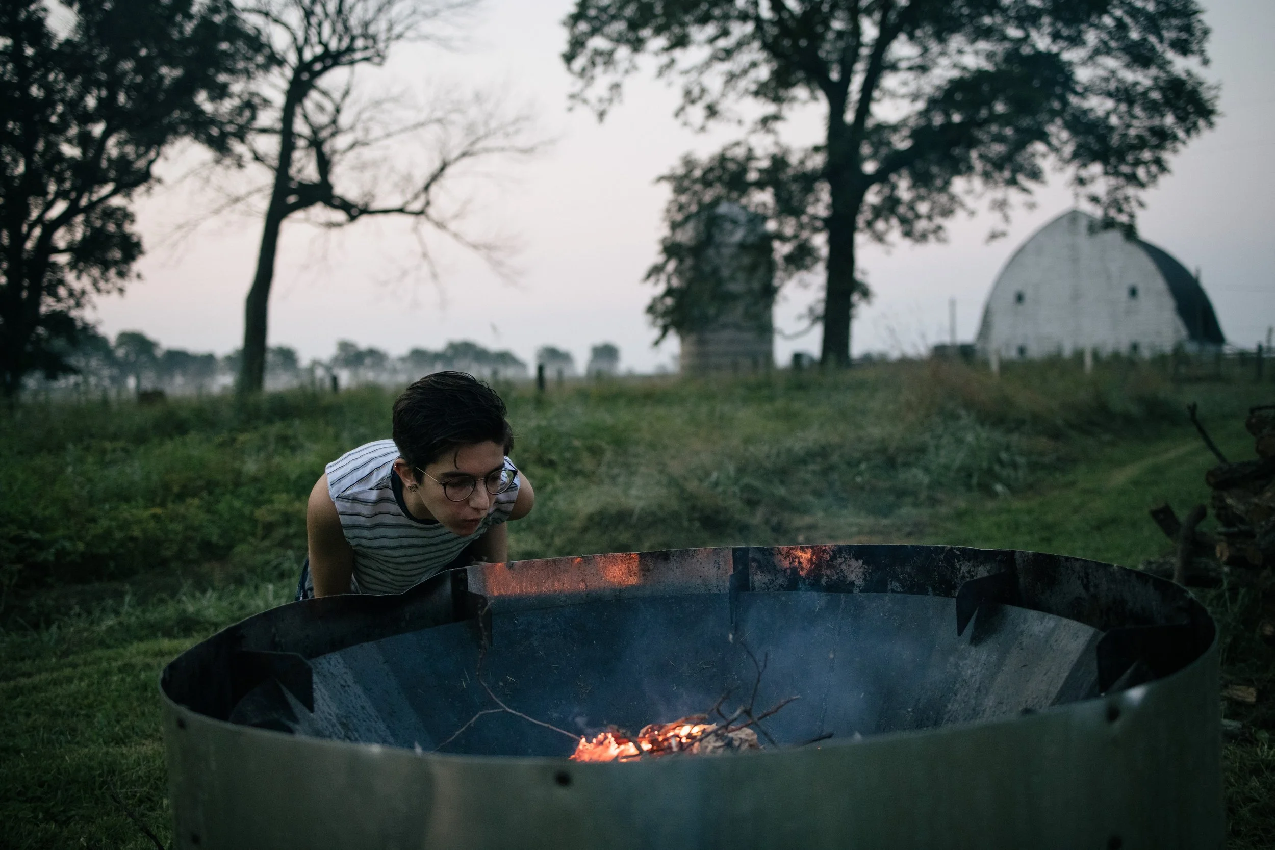 Person in striped shirt blowing on a fire pit outdoors, with trees and a barn in the background.