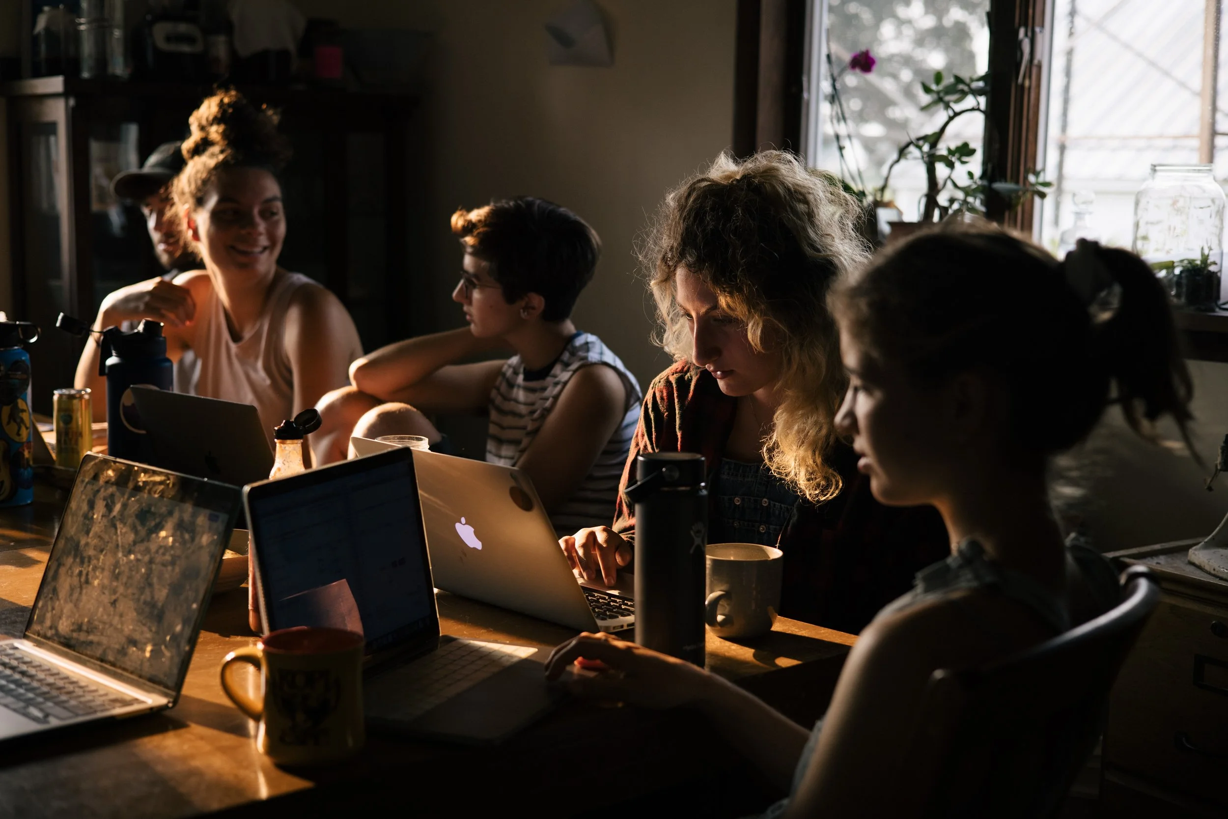 Group of young adults working on laptops around a table, with natural light streaming through a window.