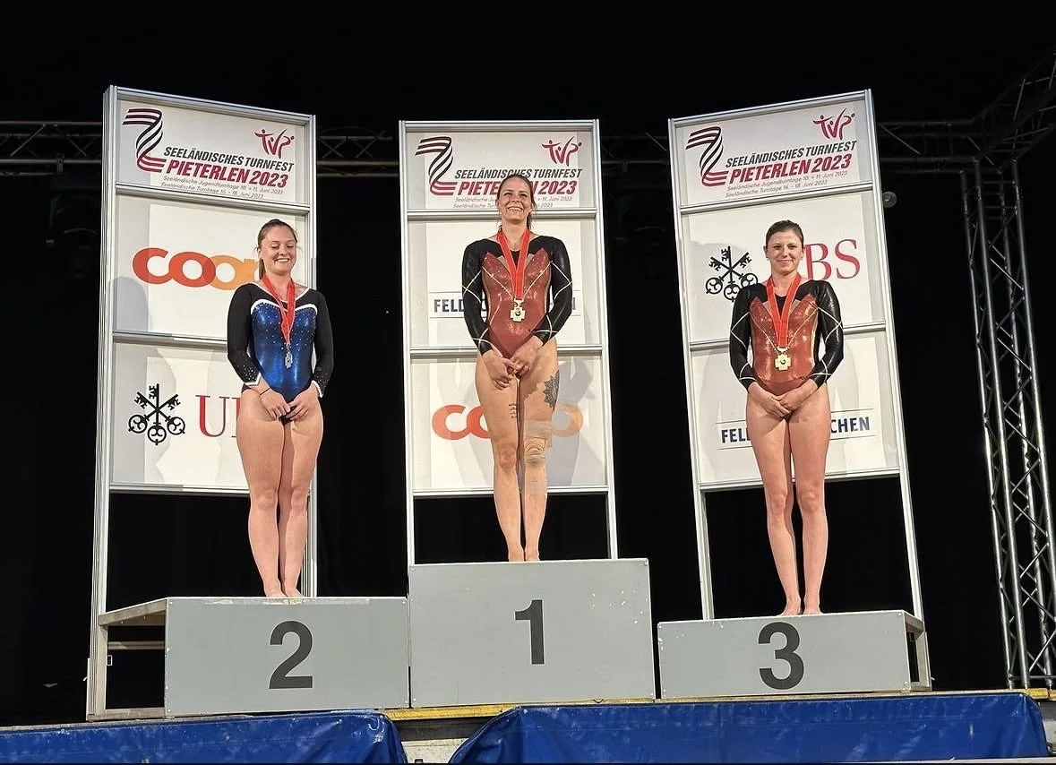 Drei Turnerinnen stehen bei einer Siegerehrung eines Geräteturnen-Wettkampfs auf dem Podest, die mittlere auf Platz 1 mit Goldmedaille und die rechte auf Platz 3 mit Bronze-Medaille sind Turnerinnen vom TV Orpund. Im Hintergrund sind Banner mit Turnf