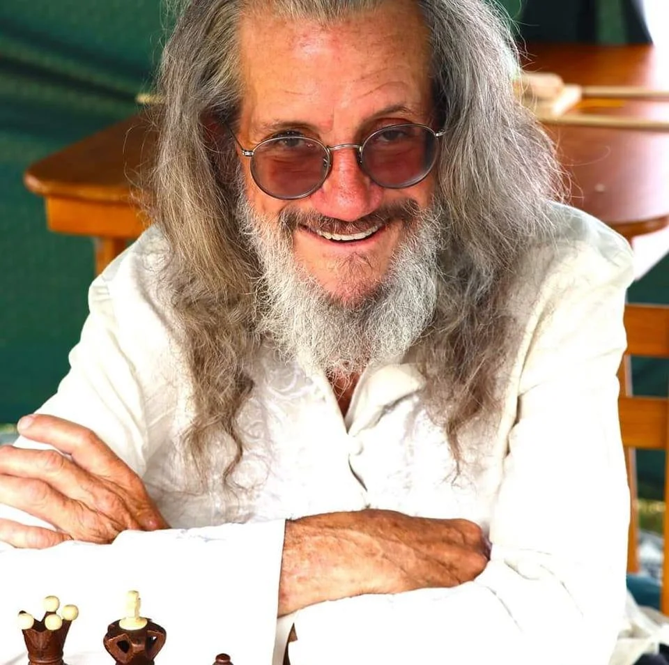 Picture of John on a white shirt wearing glasses long hair in front of a chess set